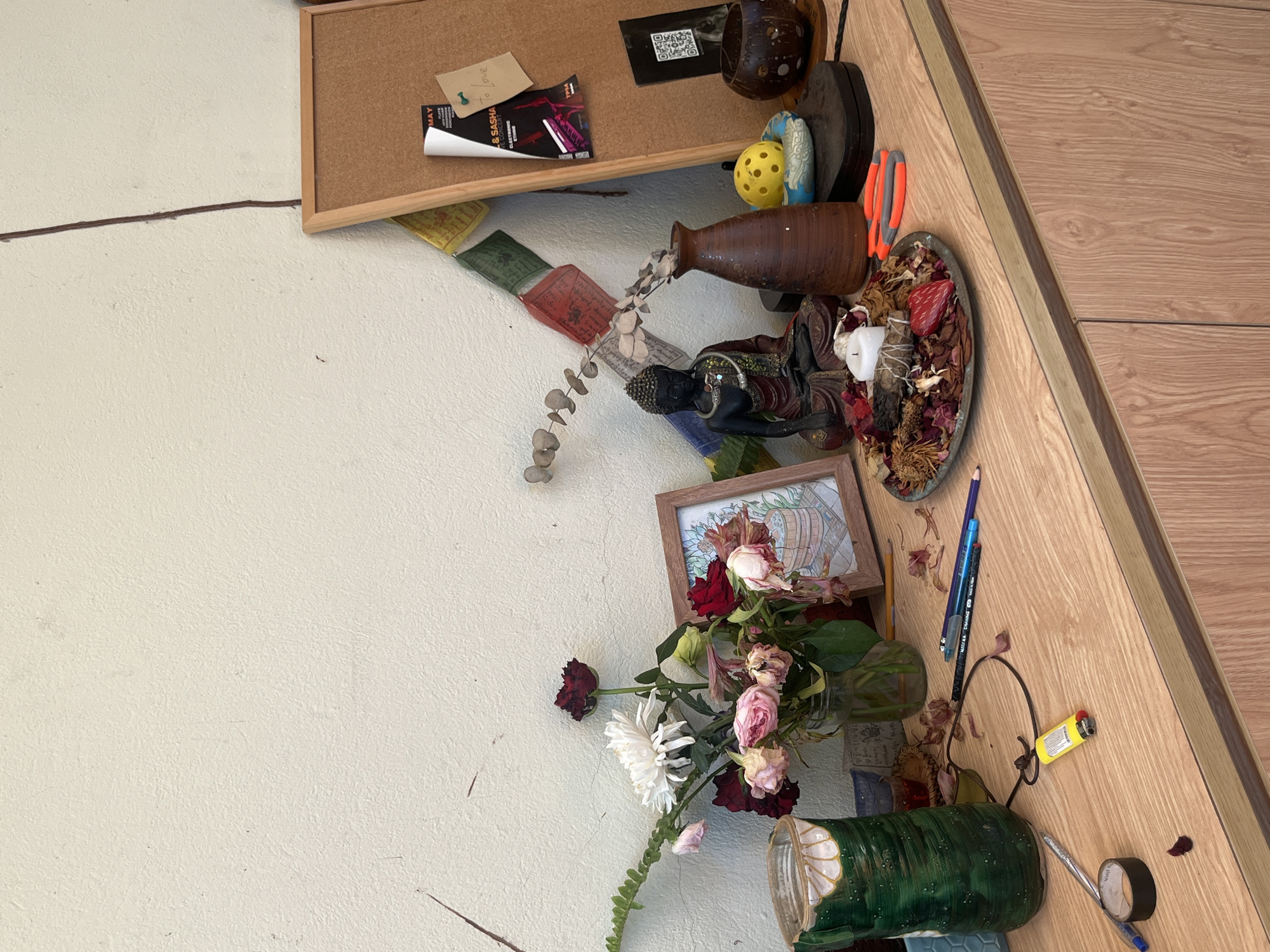 The image depicts a cluttered wooden countertop against a textured, off-white wall. The countertop displays an assortment of items, including a green vase with a decorative pattern, a jar of wilting flowers, a framed drawing of a person in a tub, a dark statue of Buddha, a brown vase, a yellow ball, a coconut shell, a circular dish filled with dried potpourri and a candle, and a pair of orange-handled scissors. Several pencils and pens are scattered on the surface, along with a yellow lighter, a roll of tape, and a blue pen. Prayer flags hang on the wall behind the Buddha statue, attached to a corkboard that also features various notes and flyers. A dark, thin line runs vertically down the wall. The scene suggests an indoor setting, possibly a desk or shelf, in Đà Nẵng, Vietnam.