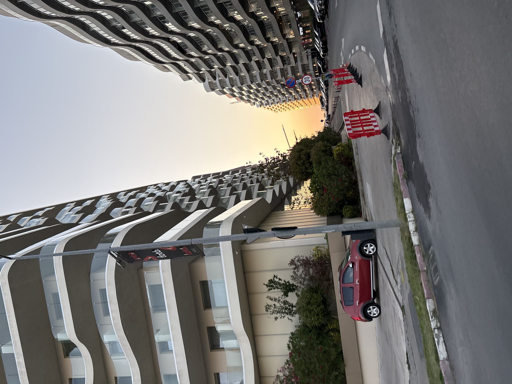 The image depicts a street scene in Năvodari, Romania, likely during sunset given the warm light. Two large, modern apartment buildings with curved balconies dominate the frame, one on the left and one on the right. The building on the left has a banner hanging from a lamppost that reads "toto". A red Dacia Sandero is parked in front of the left building. The street is paved and shows some wet patches. Red and white safety barriers are placed along the right side of the road, and several cars are parked in a line further down the street. The sky is clear and exhibits a gradient of light blue to pale orange, indicative of the setting sun. Lush green bushes and trees are visible in the foreground and between the buildings.