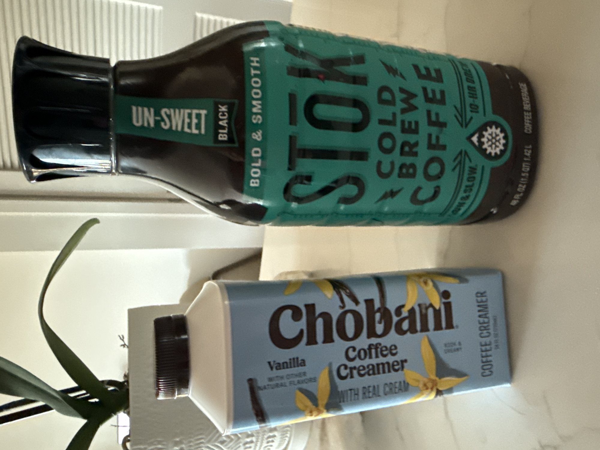 An indoor scene, likely a kitchen counter or similar surface in San Diego, United States of America, displays two beverage products and a plant. The image is oriented sideways.
On the left, a dark brown/black bottle of Stōk Cold Brew Coffee is visible. Its label, primarily teal, clearly states "STOK COLD BREW COFFEE" and identifies it as "UN-SWEET BLACK" and "BOLD & SMOOTH." Further details include "10-HR BREW," "SLOW & LOW," and "COFFEE BEVERAGE," with the volume specified as "48 FL OZ (1.5 QT) 1.42 L."
To the right of the coffee bottle, a light blue carton of Chobani Coffee Creamer is positioned. The carton features illustrations of yellow vanilla flowers and dark vanilla beans. Visible text includes "Chobani Coffee Creamer," "Vanilla," "WITH OTHER NATURAL FLAVORS," "WITH REAL CREAM," and "RICH & CREAMY." The volume is indicated as "24 FL OZ (1.5 PT) 709 mL."
Behind the Chobani creamer, a plant with long, dark green leaves, possibly an orchid, adds a decorative element. The background consists of a white, ribbed surface, suggesting blinds or cabinet doors. No human activity is depicted; the items are stationary on a light-colored surface. The overall impression is a casual setup for a coffee station.