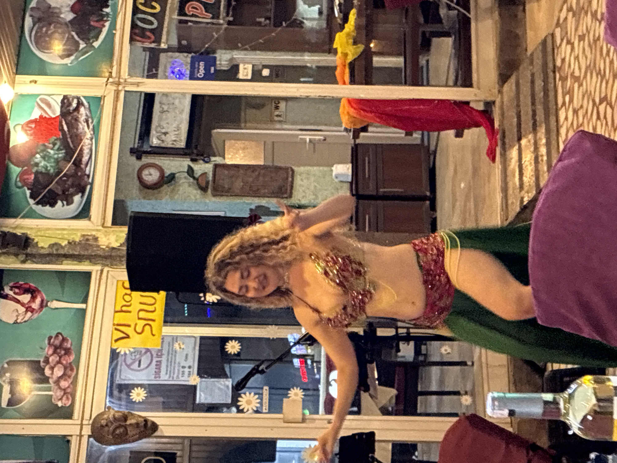 A belly dancer with curly blonde hair and a sparkling red and gold costume is performing. She is wearing a beaded bra and a matching skirt with a yellow string tied around her waist. Her eyes are closed, and she has a serene expression on her face. Her arms are outstretched, and her fingers are adorned with rings. The dancer is on a stage in front of a window display that features various items, including images of food, grapes, and a skull. The background shows a dimly lit interior with shelves and cabinets. A bottle of alcohol and other objects are visible on a table in the foreground. The scene appears to be at night, possibly in a restaurant or bar. A yellow sign with Turkish text hangs from the window, and a "No Smoking" sign is also visible. The overall atmosphere is festive and engaging.