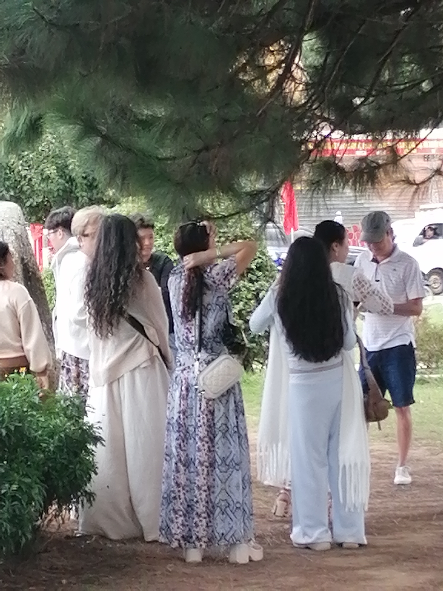 A group of individuals are gathered outdoors under the shade of pine trees, with a glimpse of buildings and a road visible in the background. The scene appears to be in Da Lat, Vietnam, suggesting a park or a public gathering space.
On the left, a woman wearing a light-colored long-sleeved shirt and pants is partially visible, her back to the camera. She is standing next to a large rock. To her right, a person with blonde hair wearing glasses and a white jacket is facing left, seemingly in conversation with another individual partially obscured.
In the center of the frame, a woman with long, dark, curly hair is wearing a light-colored cardigan over a patterned dress. She has a white crossbody bag and is touching her hair. Next to her, a person in a dark shirt is looking towards the right.
To the right of the center, a woman with long dark hair is wearing a light blue top and matching pants, draped with a white fringed scarf. She is looking towards a man wearing a white polo shirt, blue shorts, and a grey baseball cap. The man is holding a white object, possibly a paper or a small booklet, and looking down at it.
The environment is natural, with lush green foliage and the distinct texture of pine needles from the overhead trees. The ground is covered in dirt and some dry leaves. In the background, the red and yellow of some decorative banners or flags are visible, along with the side of a white vehicle. The overall impression is a casual outdoor gathering.