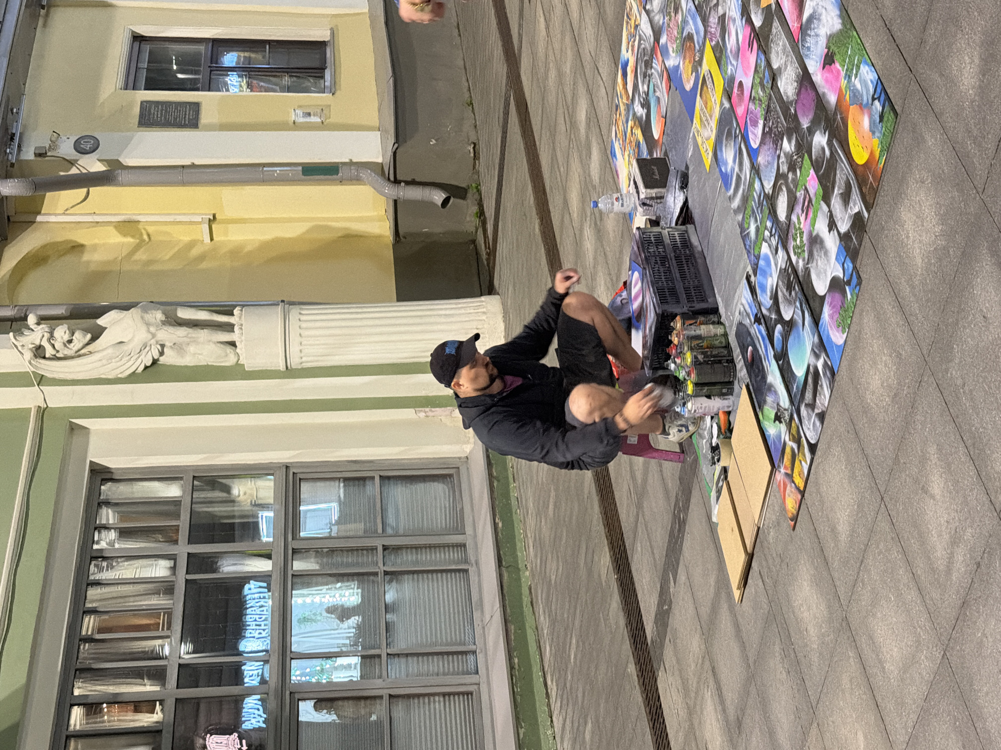 In Nizhny Novgorod, Russia, a street artist is seen squatting in front of a display of vibrant, abstract art pieces laid out on the pavement. The artist, wearing a black hooded sweatshirt and shorts, appears to be organizing or preparing spray cans, several of which are lined up on the ground next to a black crate. The artwork features swirling colors and cosmic or planetary imagery. Behind the artist, the facade of a building is visible, painted in pale green and yellow. A distinctive architectural feature, a stone sculpture of a winged creature resembling a gargoyle, stands prominently on the left. The street is paved with cobblestones, and the overall scene suggests an outdoor urban setting, possibly a marketplace or a pedestrian area where artists display their work.
