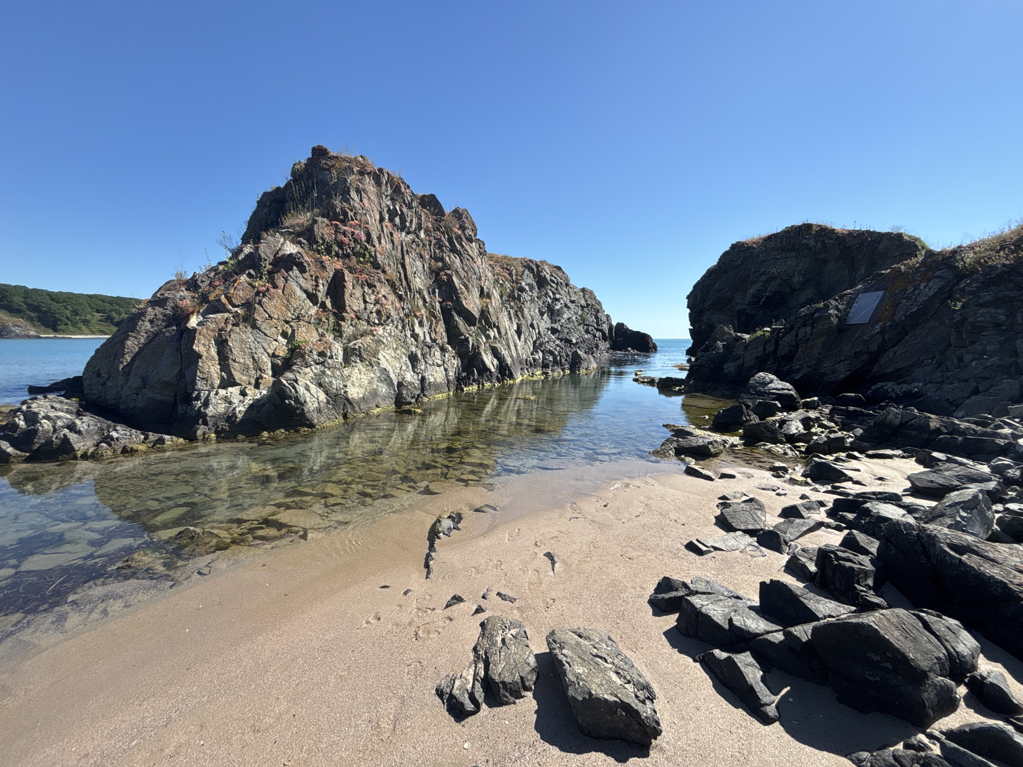This image depicts a rocky coastline near the village of Rezovo, Bulgaria, under a clear blue sky. The foreground features a sandy beach with scattered dark gray rocks, some of which are angular and others more rounded. To the left and center of the image, a large, rugged rock formation rises from the sea, characterized by its layered texture and reddish-brown and gray hues, with sparse vegetation growing on its upper slopes. The water is calm and clear, revealing submerged rocks and pebbles. Further to the right, another large, dark rock formation juts out, appearing somewhat smoother than the one on the left. A rectangular metallic plaque or sign is visible attached to the face of this rock. The background shows a distant coastline with green vegetation and the vast expanse of the blue sea meeting the horizon. The scene is devoid of any human presence or activity.