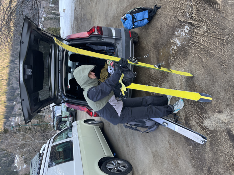 The image depicts an outdoor scene in Vallorcine, France. A single individual, dressed in an olive-green hooded jacket with black sleeves and grey mid-section, black pants, black gloves, and light-colored sneakers, is positioned facing the open rear liftgate of a dark grey SUV. The person is bent over, engaged in handling a pair of bright yellow skis. One ski is held upright, leaning against the vehicle, while its counterpart lies on the unpaved, dirty ground which displays numerous tire tracks and patches of melting snow. A black backpack rests near the individual's feet, and a blue backpack is situated further to the right on the ground. The open trunk of the SUV reveals a coiled rope and other gear. To the left, a cream and white Volkswagen van with a roof rack is parked, and a green Land Rover Defender with a roof rack is visible further in the background. The environment consists of forested mountain slopes with intermittent snow patches under natural lighting conditions.