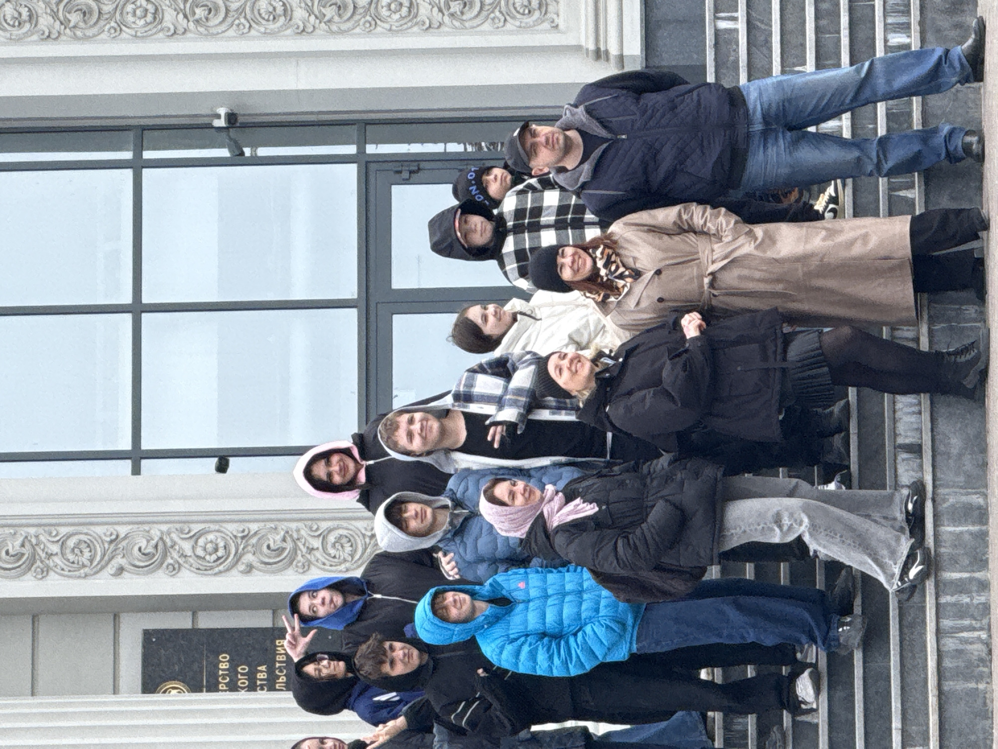 A group of approximately 14 people is standing on the steps of a building in Kazan, Russia, posing for a photo. The building has a large glass window facade and ornate architectural details above. The people are dressed in various jackets and coats, indicating a cool weather. Their expressions and postures suggest a group activity or tour. A security camera is affixed to the building above the window. A plaque with Russian text is visible on the building to the left of the group. The steps appear to be made of stone or concrete.