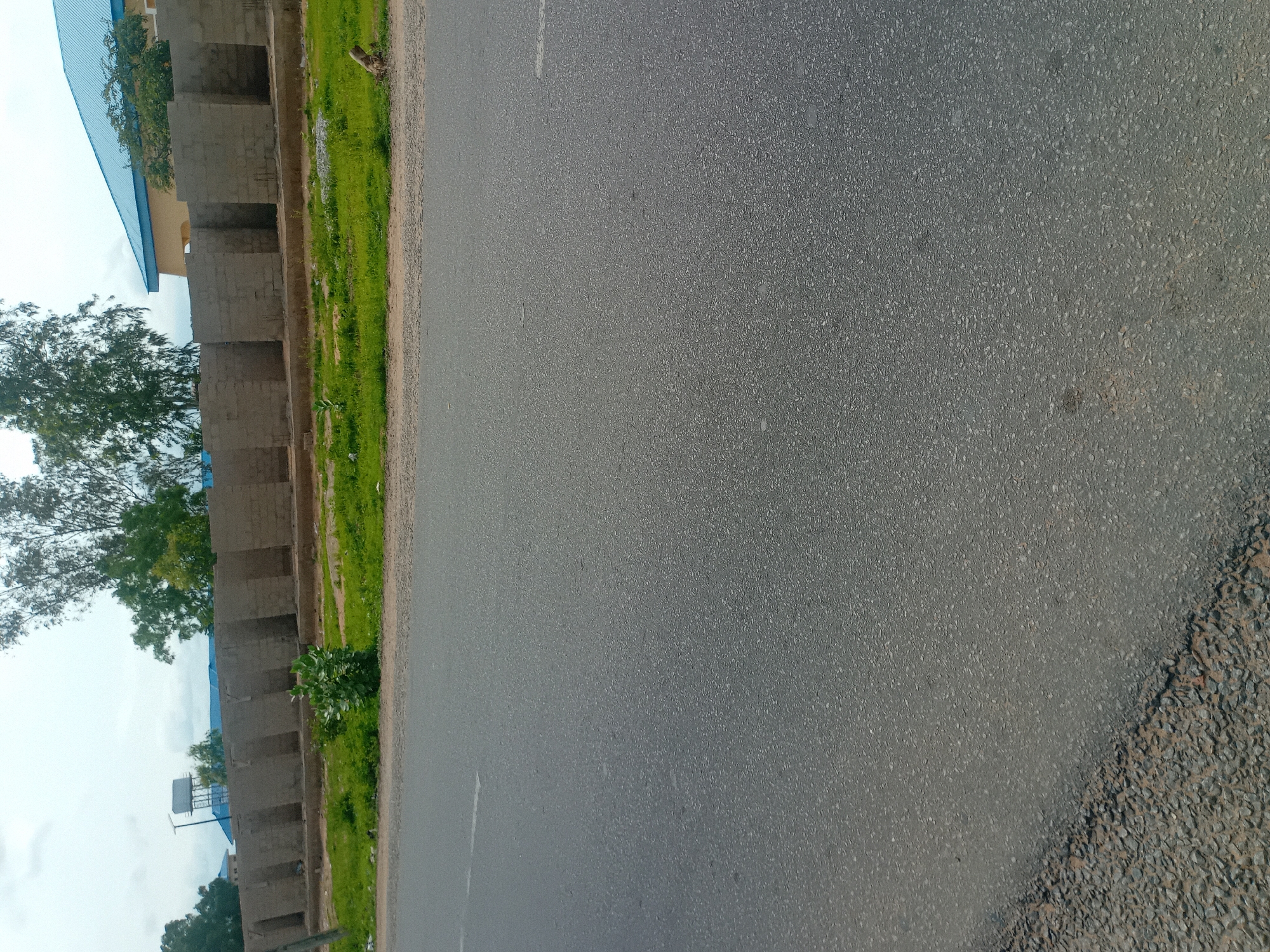 The image shows an asphalt road in Jalingo, Nigeria. A low, unfinished brick wall with regularly spaced openings runs parallel to the road, suggesting construction or development. Beyond the wall, there are trees and a building with a blue roof. The sky is overcast. There are no people or vehicles visible in the frame.