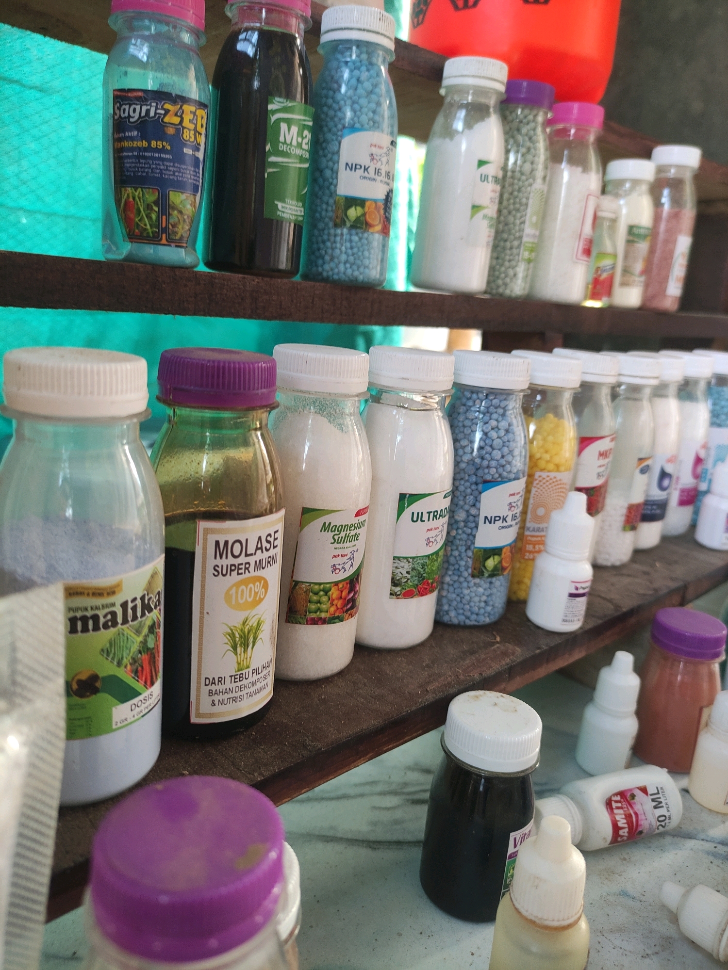 The image depicts a collection of agricultural products, likely fertilizers or pesticides, displayed on wooden shelves. The scene is consistent with a small agricultural supply store or market stall. Several bottles and containers of various sizes and colors are visible, some containing powders, granules, or liquids. Labels on some of the products are partially visible, displaying brand names such as "Sagri-ZEB", "M-2 DECOMPOSER", "ULTRA", "NPK 16.16.16", "MALIKA", "MOLASE SUPER MURNI", and "SAMITE". The background suggests an outdoor or semi-outdoor setting, with a teal-colored tarp or fabric visible on the left and the general appearance of weathered wood. The overall impression is one of a humble, functional retail space. Based on the context provided, this scene is located in the city of Tiudan, Indonesia. There are no people visible in the image.