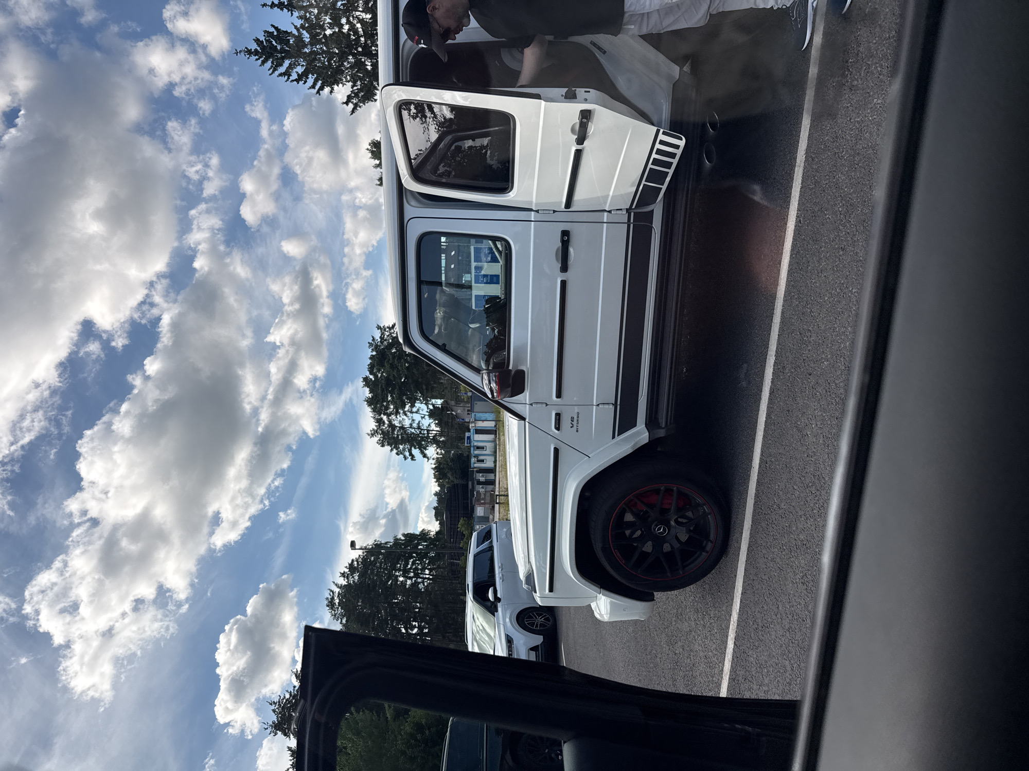 In Zhukovka, Russia, under a partly cloudy sky, a white Mercedes-Benz G-Class SUV is parked in a lot. The driver's side front door and rear door are open, with a man wearing a black t-shirt, light-colored pants, and a baseball cap exiting the vehicle from the rear passenger side. Another white SUV is parked behind it. The scene appears to be a parking lot with trees and some buildings in the background. The perspective is from inside another vehicle looking out through its open door.