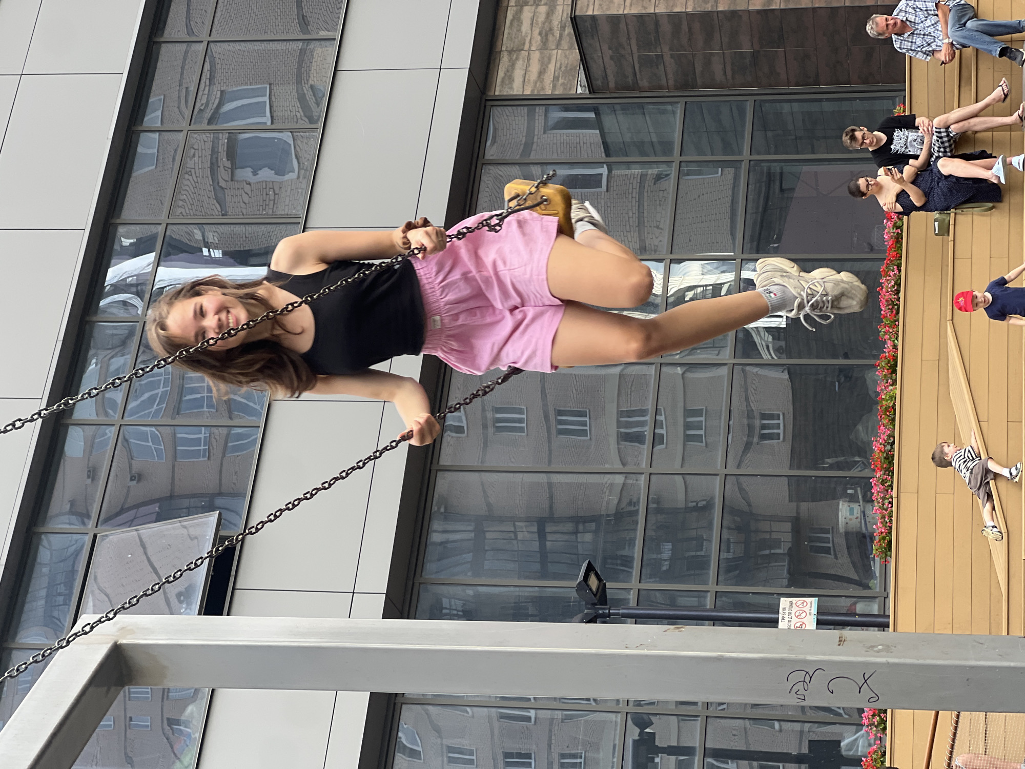 A young woman with brown hair is on a swing in an urban setting. She is wearing a black tank top and pink shorts, with white sneakers. Her legs are in motion, and she is smiling at the camera. Behind her, a modern building with reflective windows displays the distorted reflection of other buildings. In the foreground, a wooden structure with a row of pink and red flowers separates the swing area from a lower level. On this lower level, several people are sitting on a wooden bench, including a woman looking at her phone, a man with glasses, and another man. A young boy in a striped shirt and shorts is playing on a wooden ramp, and a smaller boy in a red hat is standing. The overall scene appears to be a public space in the city of Krasnoyarsk, Russia.