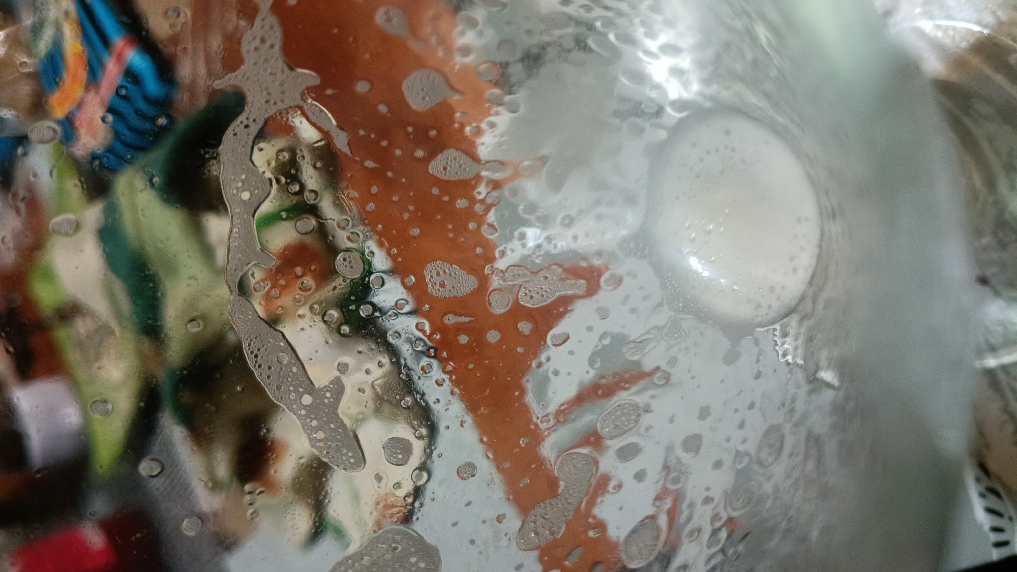 The image shows a close-up view of suds and water droplets on a glass surface, likely from inside a container or enclosure. The background is blurred and appears to show a colorful, abstract pattern with shades of orange, green, and blue. There are no discernible people or specific objects in the scene that would indicate a location in Novogusel'skii, Russia. The focus is entirely on the texture and abstract patterns created by the water and soap.