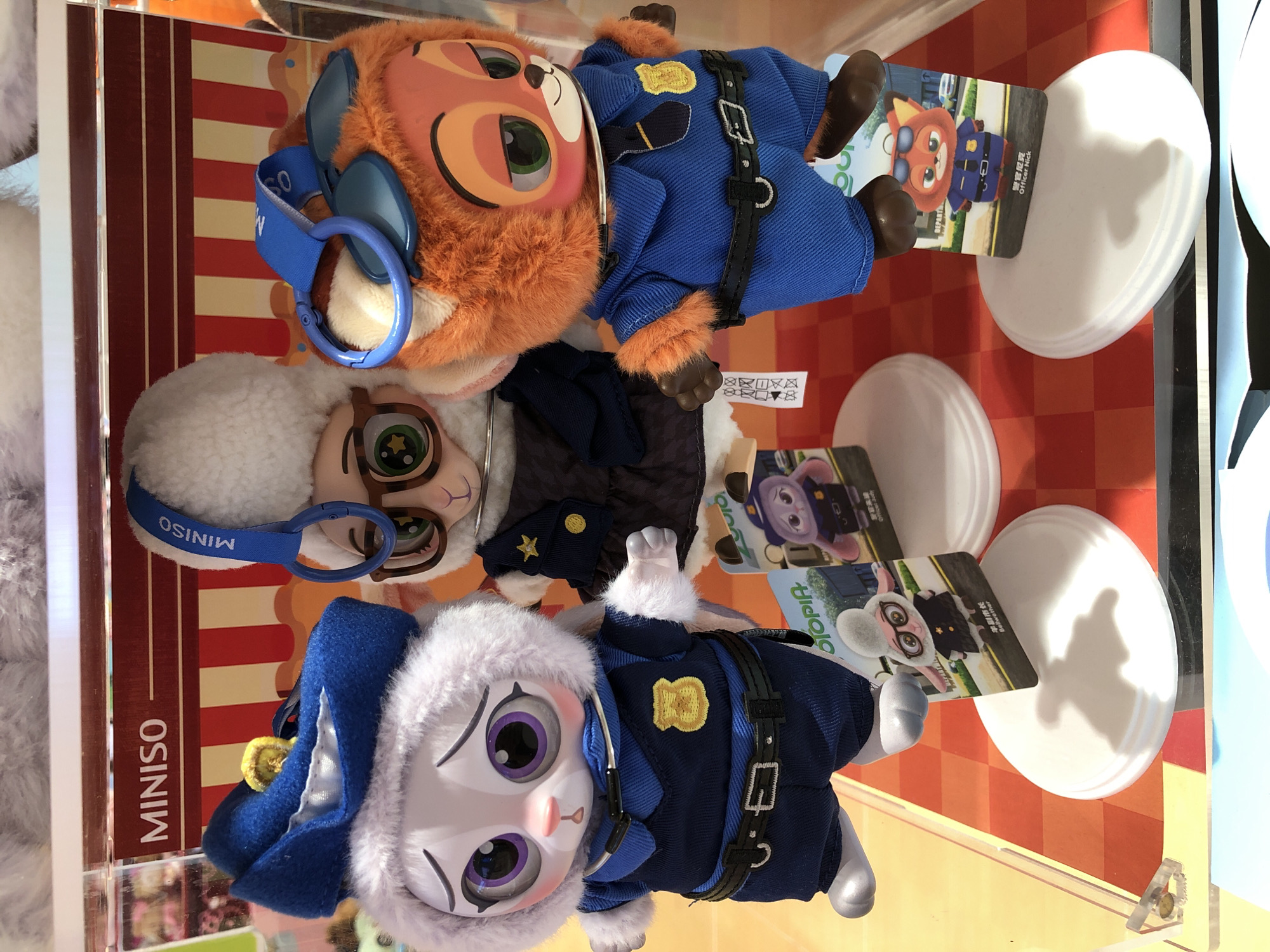 This media file shows two plush toys dressed as police officers, possibly from the Disney movie Zootopia. One plush toy is an orange fox, and the other is a white rabbit. Both are wearing blue police uniforms with black belts and badges. The fox has headphones on, and the rabbit has a police hat.
The toys are displayed on a red and white checkered surface, likely in a store or exhibit. Behind them, there is a display with red and white stripes. Visible text includes "MINISO" on a blue strap attached to the rabbit, and on a card depicting the fox, it says "警官 尼克 Officer Nick". On a card with the rabbit, it says "警官 茱蒂 Officer Judy".
The overall impression is that these are collectible toys, perhaps for sale in a store. The lighting appears to be indoor, and there are no indications of the time of day or weather. The scene is well-lit, highlighting the details of the toys. There is no specific emotion displayed by the toys.