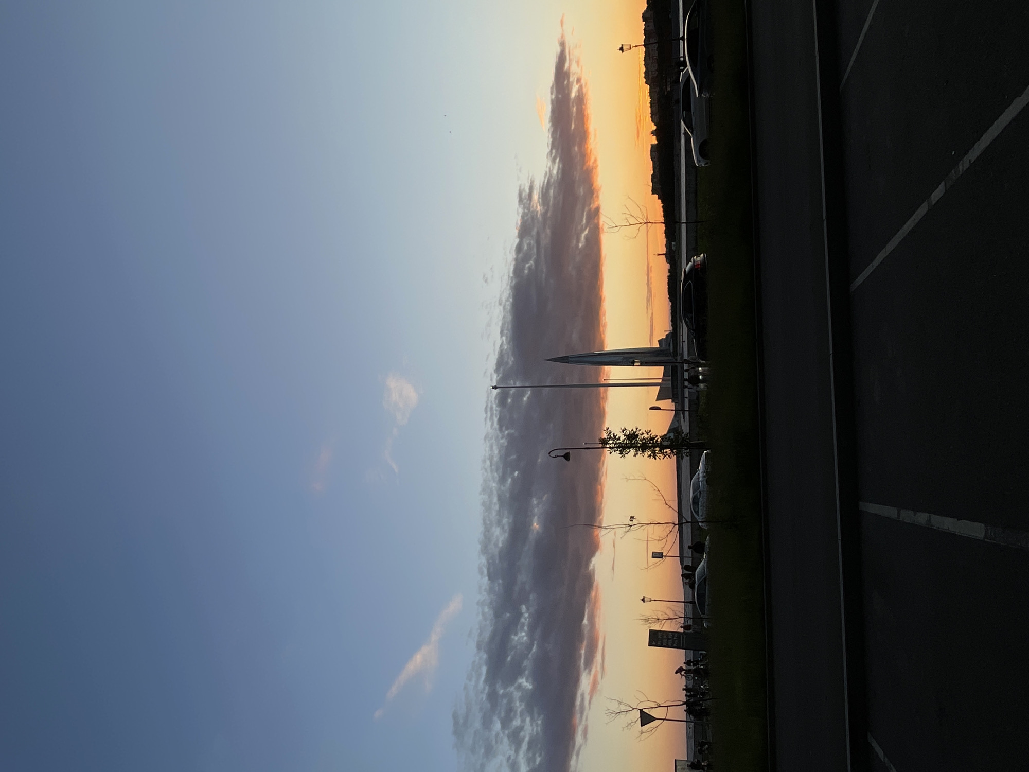 A vertical, wide-angle shot captures a city park and waterfront at sunset. The sky transitions from a light blue at the top to an orange and pink gradient near the horizon, highlighted by a long, dark cloud formation. In the midground, a line of silhouetted people and bicycles are gathered along a waterfront path, some near informational signs. Several cars are parked on a grassy strip bordering the path, with bare trees interspersed. Prominent structures include a tall, pointed spire on the left and a building with a domed roof on the right. The foreground shows a dark asphalt surface with white parking lines. The overall scene depicts a public space experiencing the end of daylight.