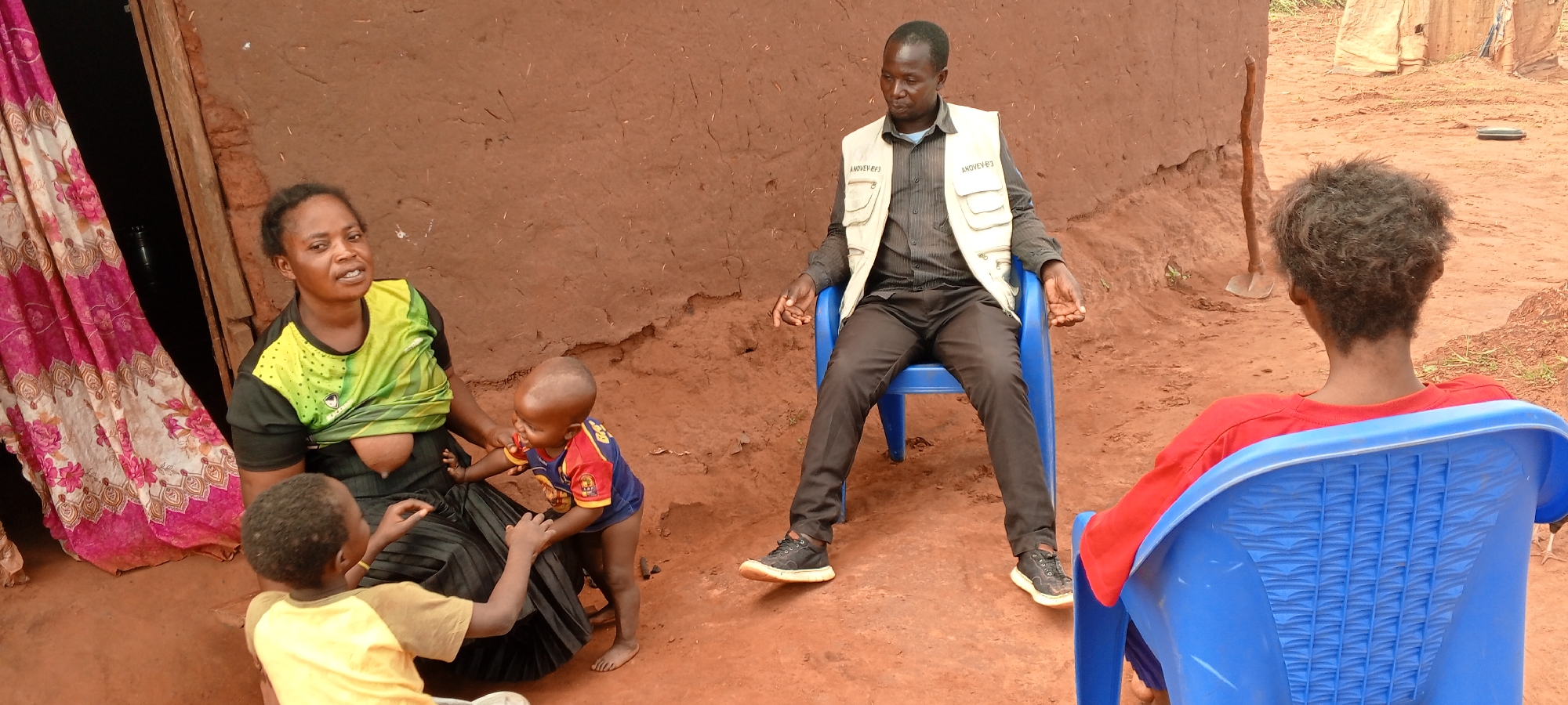 The image captures a scene in the city of Makungu, Democratic Republic of the Congo. A woman is seated near a doorway covered by a floral curtain, breastfeeding her child. Two other young children are near her, one touching her. A man wearing a vest with the word "ANOVEF/3" is seated on a blue plastic chair, facing the group. Another person, wearing a red shirt and seated on a blue plastic chair, is facing away from the camera. The background consists of a mud wall and a dirt ground.