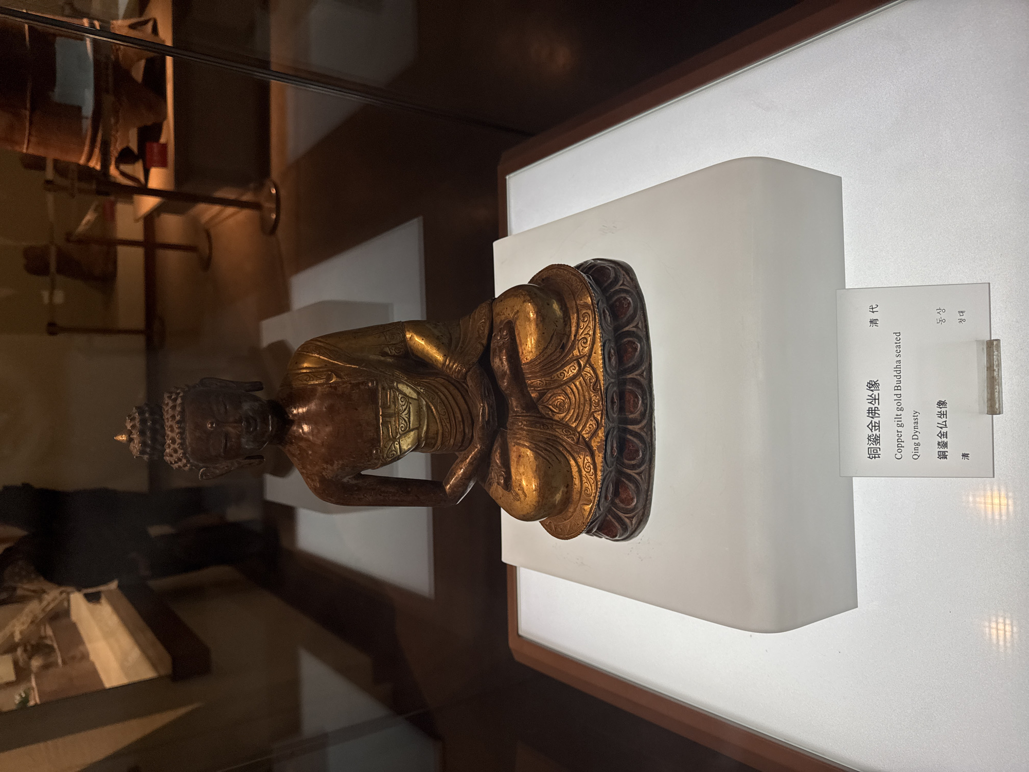 The image shows a copper gilt gold Buddha seated statue, identified as being from the Qing Dynasty. It is displayed on a white pedestal inside a museum exhibit. The information card is visible, providing the name of the artifact in multiple languages: Chinese, English, Japanese, and Korean. The statue is a golden hue with detailed carvings, and the exhibit case shows reflections of other objects in the museum, suggesting it is enclosed in glass. The statue is placed within an art museum or similar cultural institution in Dunhuang City, China.