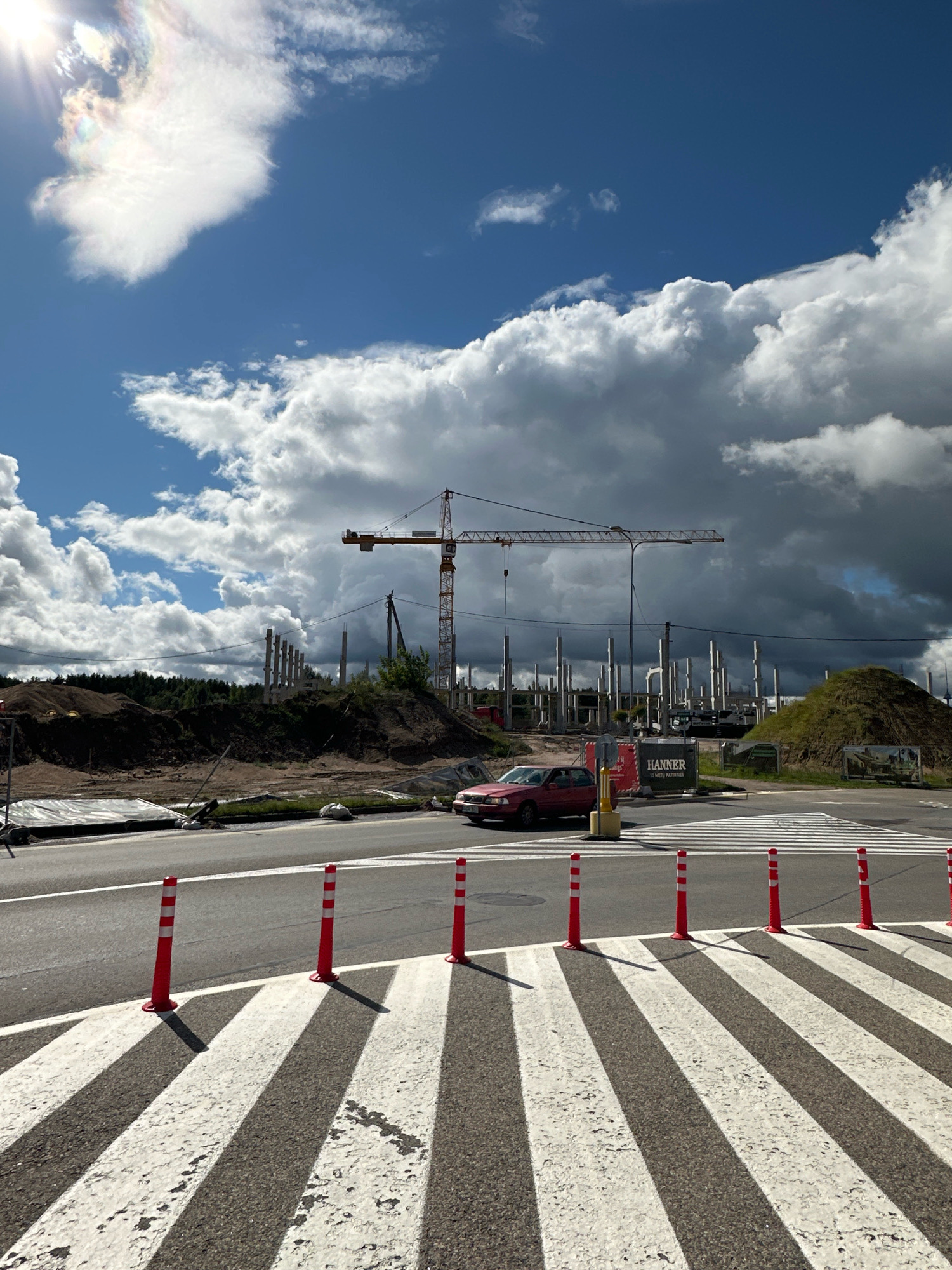 This is a photograph of a construction site in Vilnius, Lithuania. The foreground shows a pedestrian crossing with white and black stripes, bordered by red and white traffic cones. A red car is stopped on the road near the crossing. In the background, a large yellow construction crane is visible, along with numerous concrete pillars that appear to be part of a building under construction. The site is situated on a hill with piles of dirt. The sky is blue with dramatic, dark clouds, and the sun is shining brightly, creating a rainbow-like effect in the upper left corner.