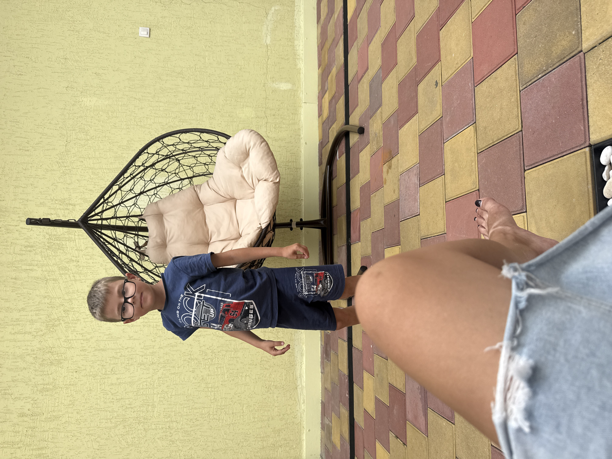 In Sirius, Russia, a young boy wearing glasses, a blue t-shirt, and blue shorts stands in front of a pale yellow textured wall. He is positioned to the left of a black hanging chair with a beige cushion. The ground is paved with square interlocking tiles in shades of red and yellow. The perspective suggests the photo is taken from a low angle, with a person's legs and denim shorts visible in the foreground, partially obscuring the tiled ground and the lower portion of the scene.
