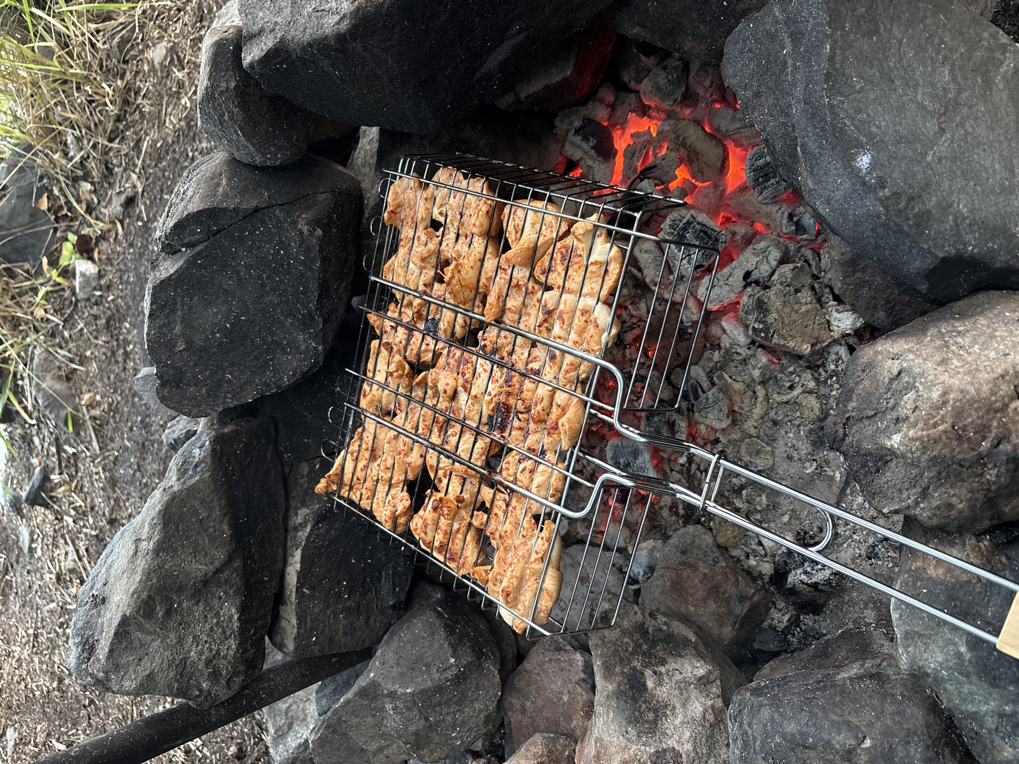 The image depicts a close-up, elevated view of chicken pieces being grilled on a metal basket over glowing red embers in a fire pit. The fire pit is constructed from large, dark grey rocks. The ground around the fire pit appears to be composed of dirt and dry vegetation. The scene is set outdoors, likely in a natural, undeveloped area given the presence of rocks and wild grass. The overall impression is one of an outdoor cooking activity.
