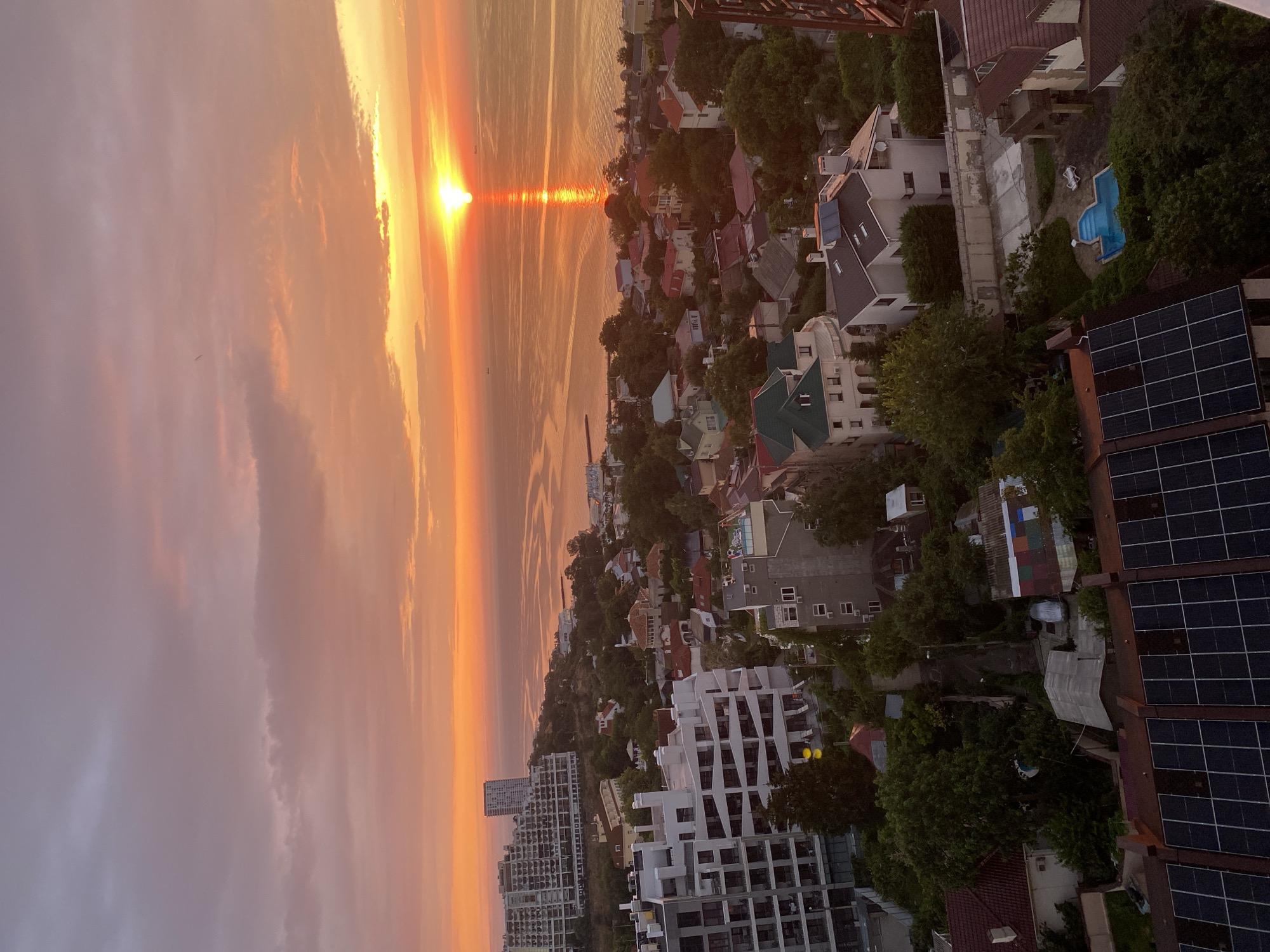 An aerial view captures a vibrant sunset over the city of Odesa, Ukraine. The sky is a canvas of warm oranges, pinks, and purples, with the sun casting a shimmering reflection across the calm sea. In the foreground, a dense urban landscape unfolds, characterized by a mix of residential buildings and modern high-rises. Lush green trees are interspersed throughout the neighborhood, softening the architectural edges. The scene includes a prominent building with a distinctive layered facade, several multi-story apartment complexes, and smaller houses with red-tiled roofs. A small, inviting swimming pool with lounge chairs is visible in one of the courtyards. Solar panels are installed on the roofs of some buildings, indicating a blend of traditional and contemporary features within the city. No people are visible in the image.
