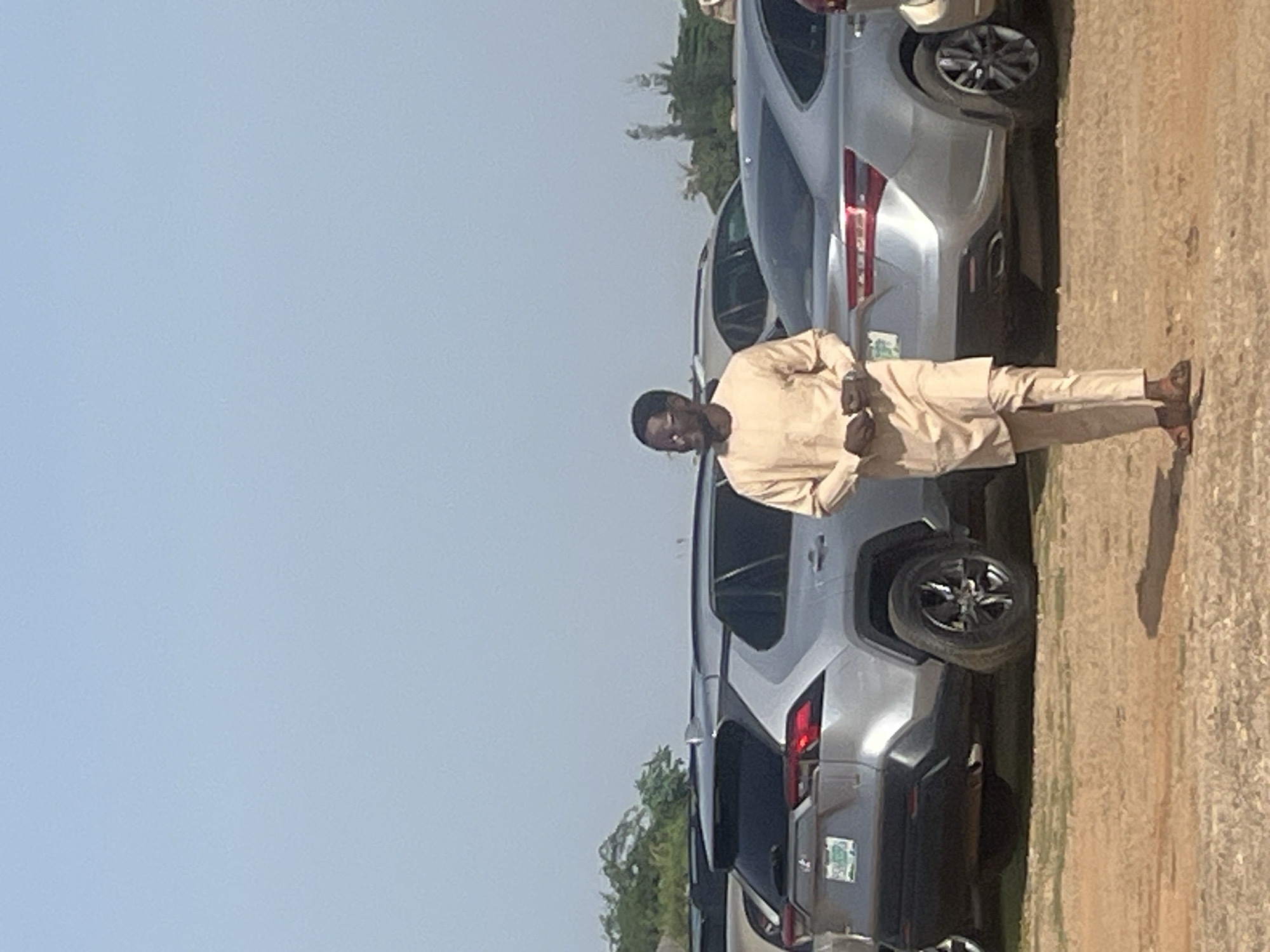 A man stands in a parking lot in Galadimawa Village, Nigeria. He is wearing a light-colored, traditional outfit and sandals. Several silver-colored SUVs are parked behind him. The sky is clear and blue. The ground is dirt or gravel.