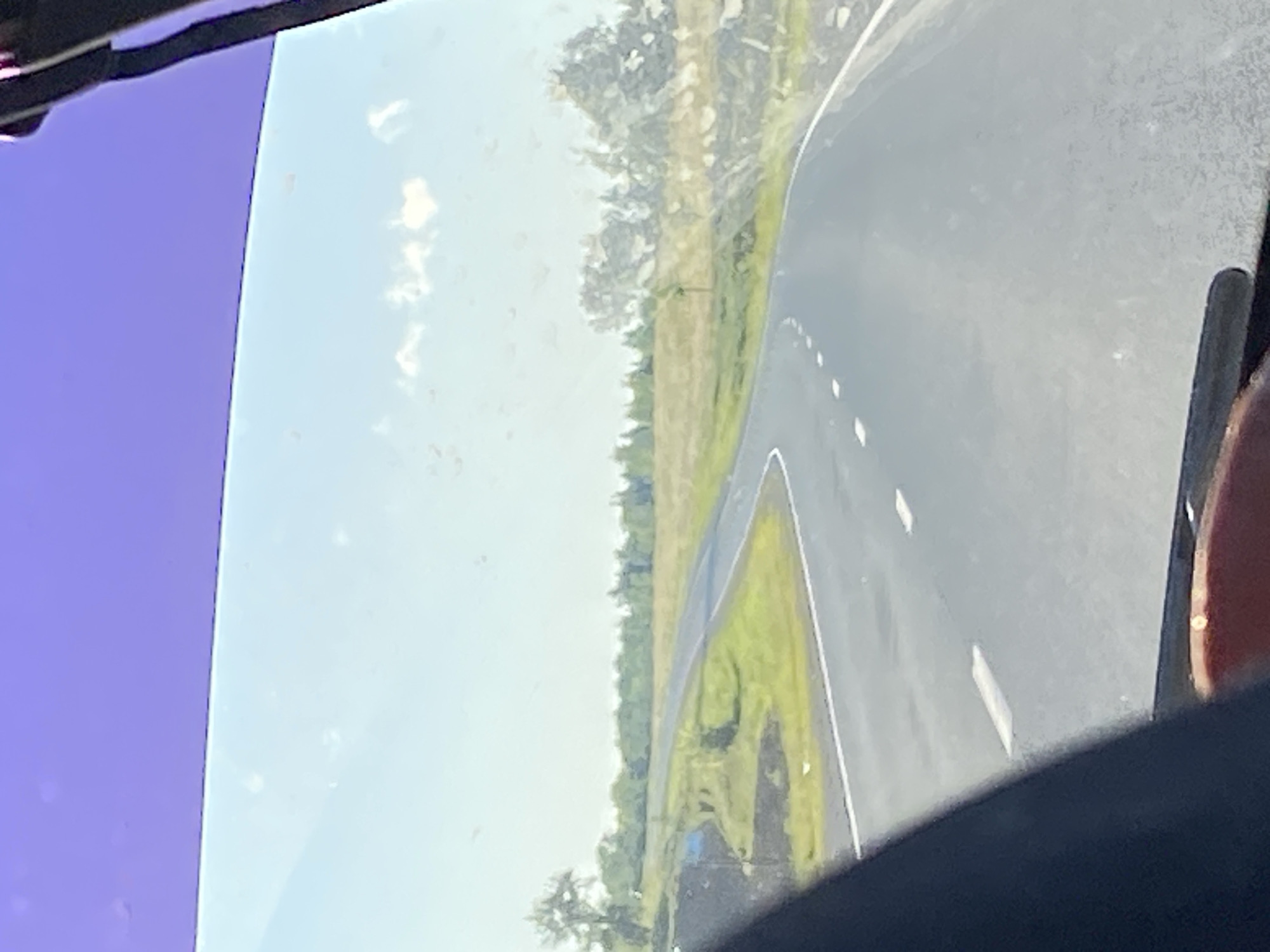 The image shows a view from inside a vehicle looking out through a windshield onto a two-lane asphalt road that curves to the left. The road has white dashed lines marking the lanes. On either side of the road are grassy fields with scattered trees and bushes. In the distance, a line of trees forms a horizon. The sky is clear with a few wispy clouds. The upper portion of the windshield has a purple tint. The foreground of the image is dominated by a dark, blurry shape, likely the interior of the vehicle, with a red object visible at the bottom. The scene is outdoors, likely during daytime, given the bright lighting. The location is identified as Заглядовка, Russia.