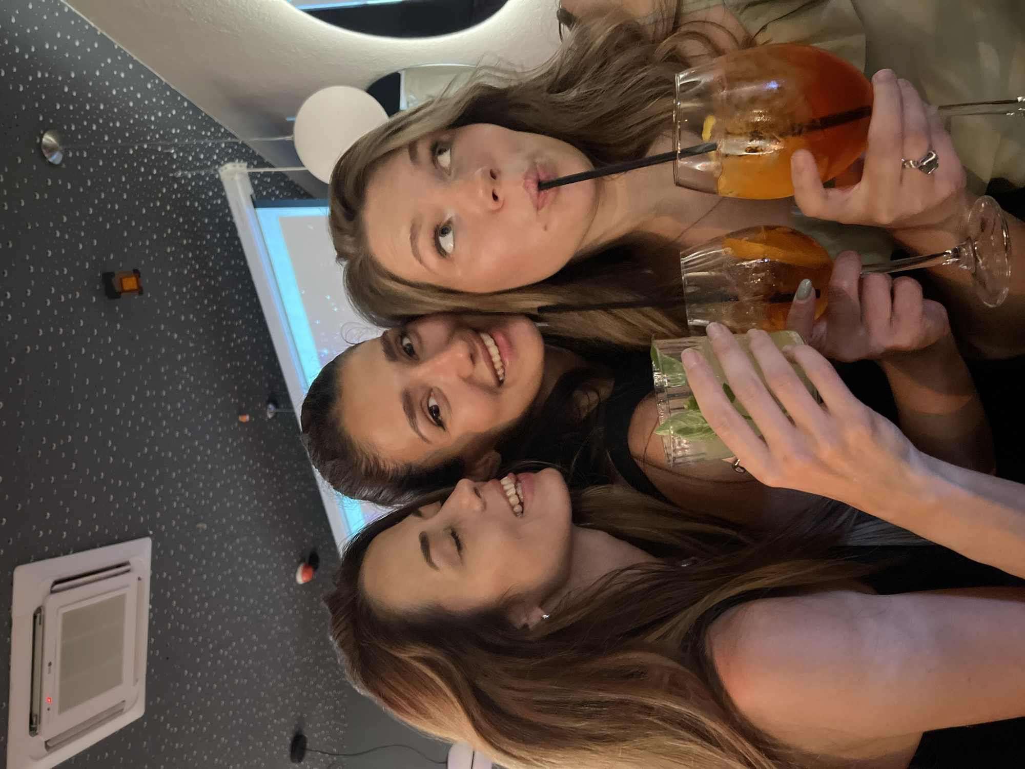 Three adult females are depicted indoors, holding various alcoholic beverages, in a social setting located in Rafailovići, Montenegro.
**Individuals:**
* **Left Female:** Possesses long, straight, dark brown hair. She is shown in profile, looking to her right, smiling broadly with visible teeth, and her eyes partially closed. She wears a dark-colored sleeveless top. Her right hand is visible, holding a clear stemmed glass containing a light green liquid and mint leaves. A small silver hoop earring is present in her left ear.
* **Middle Female:** Has dark hair pulled back from her face. She is smiling directly forward, with visible teeth. She wears a dark-colored top. Her left hand holds a clear stemmed glass containing a light green liquid and mint leaves, identical to the drink held by the left female.
* **Right Female:** Features wavy, medium-length light brown hair. She is looking upwards and to her right, with her lips pursed around a black drinking straw. She wears a light-colored top with a ruffled texture. Her right hand holds a large, stemmed balloon glass containing an orange-red liquid, an orange slice, and a black straw. A silver-toned ring is worn on her left ring finger. Her left hand holds a second identical balloon glass.
**Beverages:** Four cocktails are visible. Two are in clear stemmed glasses, containing a light green liquid garnished with mint leaves. The other two are in large stemmed balloon glasses, containing an orange-red liquid with an orange slice and a black straw.
**Environment and Decor:**
* **Ceiling:** The ceiling is dark, likely black or dark grey, and covered with a uniform pattern of numerous small, irregularly shaped white dots.
* **Ceiling Fixtures:** A white, square, ceiling-mounted air conditioning unit with a red indicator light is visible in the upper left quadrant. Several small, circular, recessed light fixtures are distributed across the ceiling. A small, rectangular, orange-brown object is mounted on the ceiling in the mid-left. Black electrical cables for pendant lights are also present.
* **Background Elements:** In the upper right background, a large white projection screen or wall displays a light blue and white abstract image, possibly depicting stars or a cosmic scene. A spherical white pendant light fixture hangs to the right of this screen. To the far right, a vertical, ribbed, white sculptural element is partially visible.
* **Lighting:** The scene is illuminated by ambient artificial light sources, creating soft shadows.