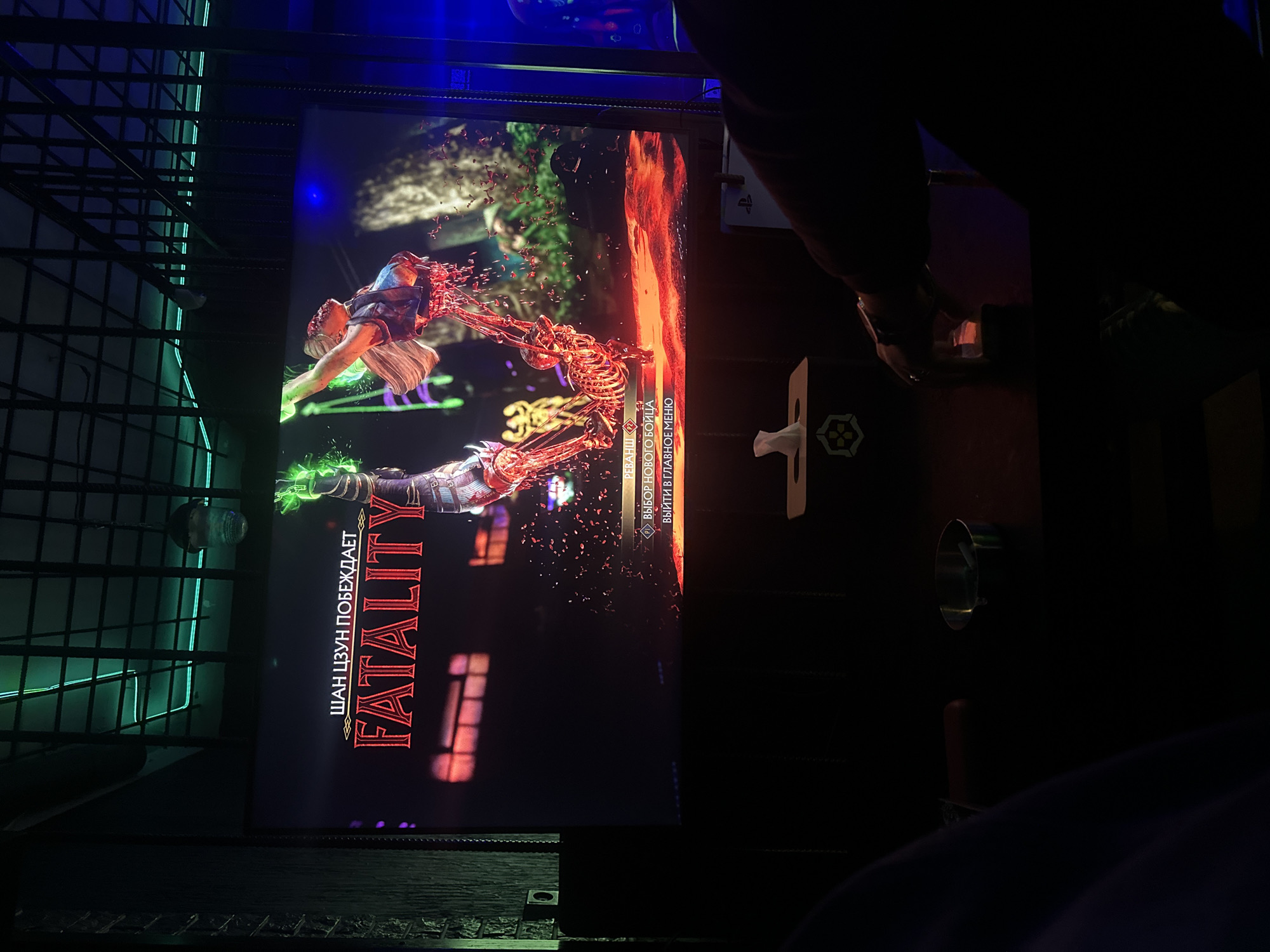 A person is playing Mortal Kombat on a screen in what appears to be an arcade or gaming room. The screen displays the word "FATALITY" in Russian along the left side. A character is performing a fatal move, with blood and skeletal details visible on the screen. A tissue box with a hexagonal logo sits to the right of the screen, next to a cylindrical metallic object. A black wire mesh structure surrounds the screen on the left, and is lit with green neon lights. The location may be in Yerevan, Armenia, though this cannot be verified from the image.
