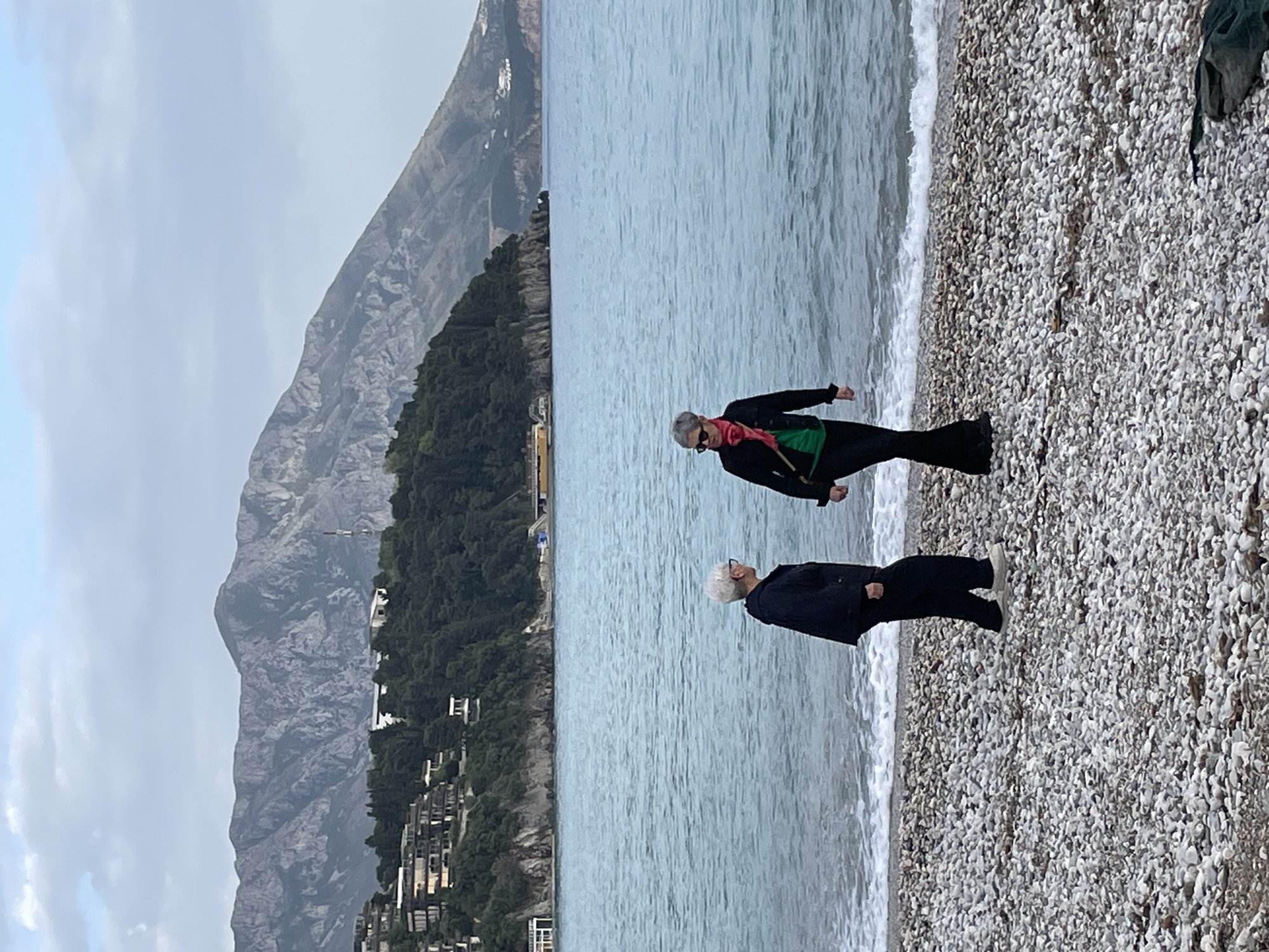 The image shows two people standing on a pebble beach in Budva, Montenegro. The beach is lined with grey and white pebbles, with the sea on one side and a mountainside with buildings and trees on the other. The sky is overcast.
The person closer to the camera is wearing a dark jacket, pants, and sneakers. The person further away is wearing a dark jacket, black pants, a green shirt, a pink scarf, and sunglasses. They are looking in the same direction.