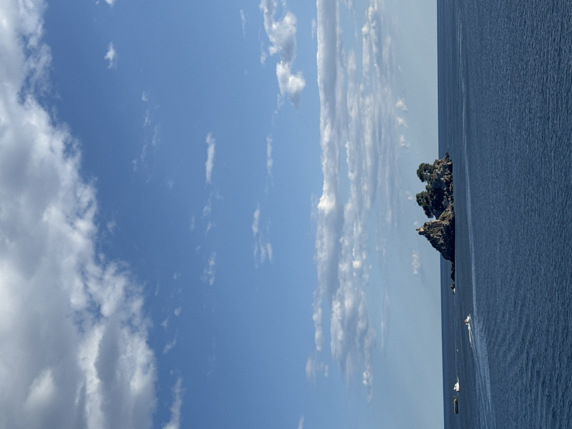 In the city of Petrovac centar, Montenegro, a body of deep blue water stretches to the horizon under a bright blue sky dotted with white, wispy clouds. In the distance, a small, rocky island rises from the sea, crowned with a cluster of trees. Atop the island, a small building, possibly a lighthouse, is visible. Several boats navigate the water: a white boat with a wake is moving away from the viewer, while another, longer white vessel is further to the left, also moving away. A dark, rocky outcrop is situated to the left of the main island. The overall scene is calm and serene, with the vastness of the sea and sky dominating the composition.