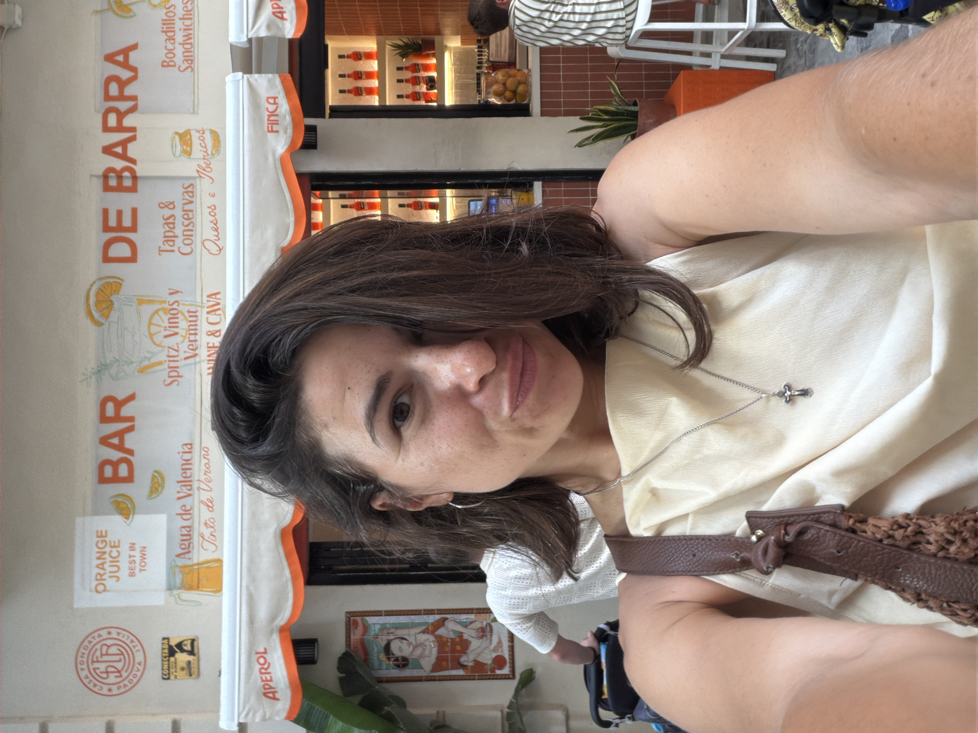 A woman with dark brown hair is taking a selfie in front of a bar. She is wearing a light-colored dress and a brown purse. The bar has a sign that says "BAR DE BARRA" and advertises various drinks and food items. There are bottles of Aperol visible on shelves behind the bar, and an orange pitcher with a straw is depicted on the sign. In the background, another person in a white shirt is visible, and a woman in a striped shirt is sitting on a stool at the bar. The environment appears to be an outdoor cafe or bar.