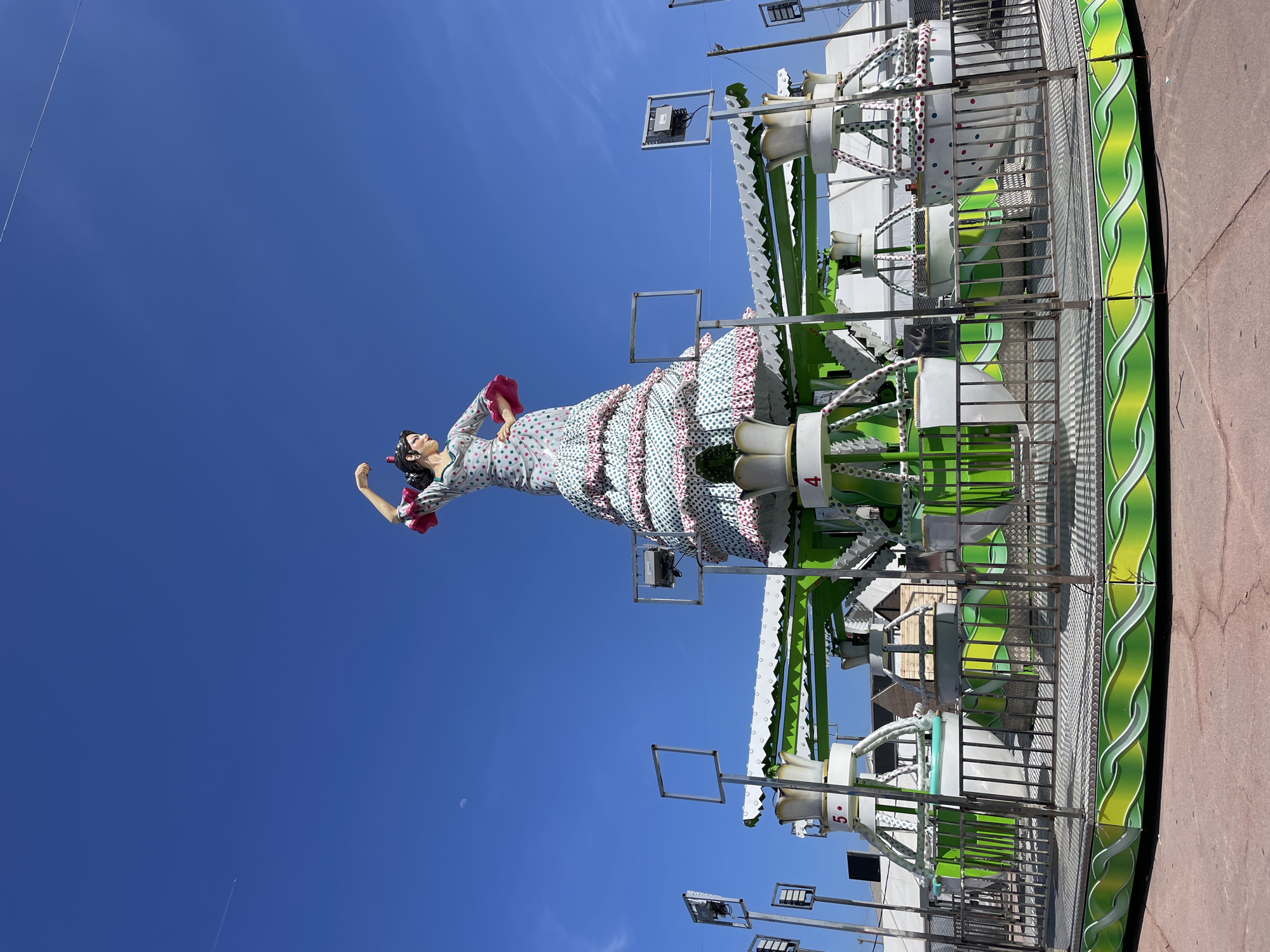 An outdoor, brightly lit scene in Sant Adrià de Besòs, Spain, depicts a large, colorful statue of a woman standing upright against a clear blue sky. The statue, reminiscent of a flamenco dancer or "gitana" figure, wears a long, flowing dress with a light blue or white base adorned with green and pink polka dots and ruffled pink trim. Her dark hair is styled up, and she holds a pink object, possibly a fan, in one hand.
Behind and to the right of the statue, a fairground ride structure is visible. It features multiple white and green elements, including what appear to be seating gondolas or pods, some of which display numbers like "4" and "5." The ride has metal railings and various technological components, including square metal frames that might hold lights or speakers, and actual speaker horns are attached to the structure. The ground beneath the ride is a reddish-brown pavement or asphalt.
The image captures a sunny day with a pristine blue sky, where a faint crescent moon is also visible in the distance, along with a contrail from an airplane. The overall atmosphere suggests a lively and festive environment typical of an amusement park or local fair.