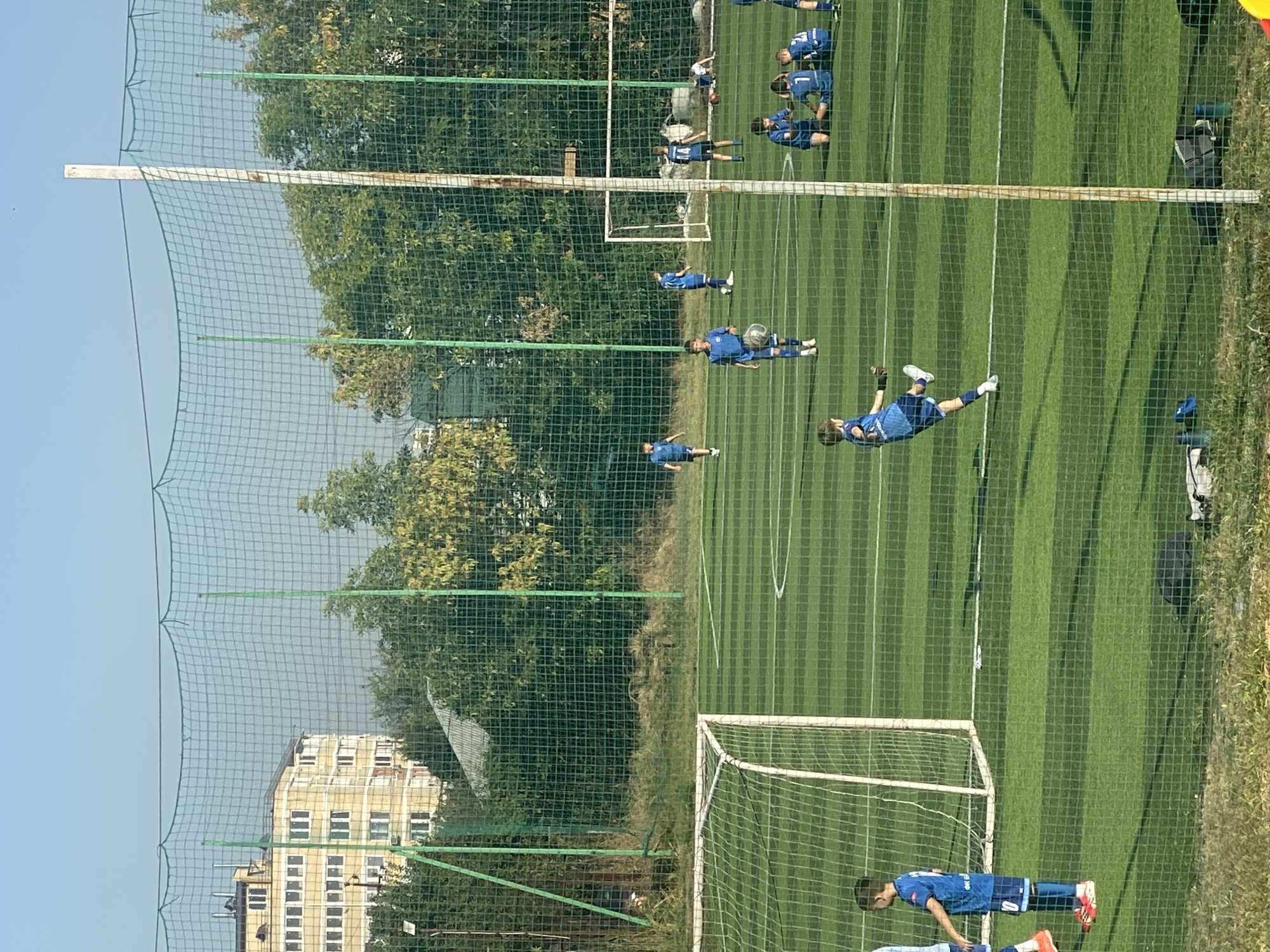 The image depicts a youth soccer practice session taking place on an artificial turf field. The field is lined with white markings and enclosed by a green net fence. In the background, a light yellow, multi-story building with numerous windows is visible, surrounded by trees. Several young boys, dressed in blue soccer uniforms, are engaged in various activities on the field, including kicking a soccer ball, running, and sitting. One boy in the foreground is actively kicking a soccer ball. Two soccer goals are present on the field. The overall environment suggests an outdoor recreational facility within an urban setting. The location is identified as Krasnodar, Russia.