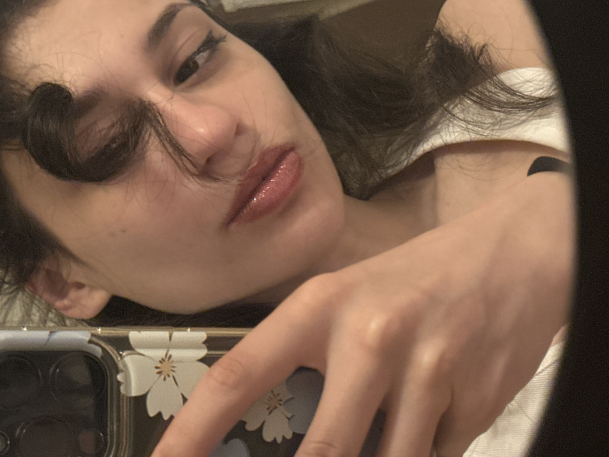 A close-up, overhead selfie of a young woman is captured against a neutral background. The woman, with dark hair and light skin, is looking downwards, with a strand of hair obscuring part of her face. She is wearing a white top and holding a phone with a floral pattern in her left hand. The image is sharp and well-lit, with no visible signs of degradation. The context of the location in Sofia, Bulgaria, is not discernible from the visual information provided in the image.