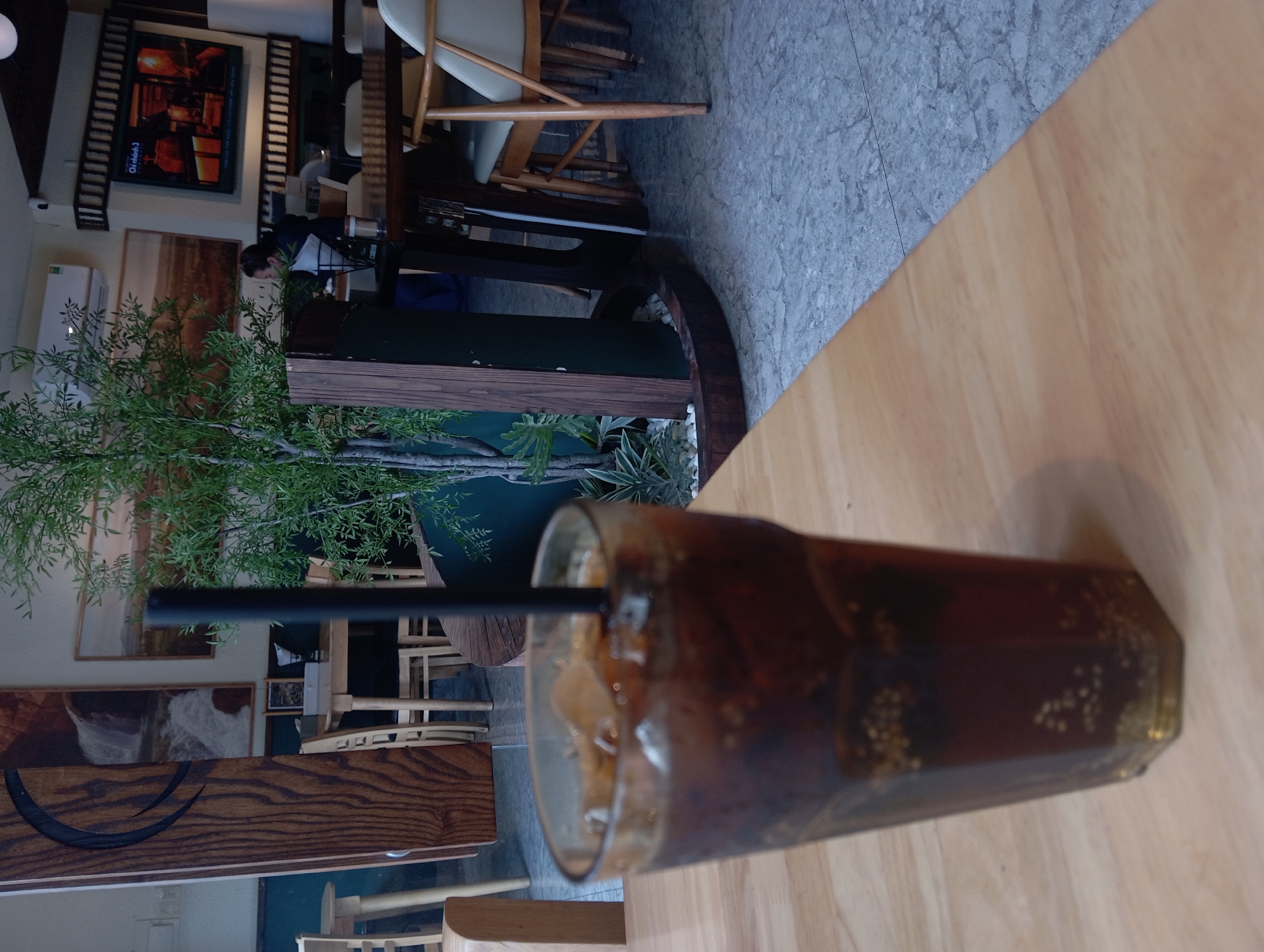 This is a medium shot image taken from a slightly low angle inside a cafe, likely in Nha Trang, Vietnam, given the context of the image's origin. The foreground is dominated by a tall glass of cola with ice and a black straw, placed on a light wood table. The glass is hexagonal and partially filled with a dark brown liquid and ice cubes, with small bubbles visible throughout.
Behind the glass, the cafe's interior is visible. To the left, a tall, dark wood panel with a carved dark circular design is present. Further back, several light wood tables and chairs are arranged. One table has a white box on it. In the middle ground, a decorative plant with green foliage is housed in a dark brown planter. A black, vertical pillar is positioned in front of the plant.
To the right, in the background, a person is seated at a table, facing away from the camera and partially obscured by a plant and a pillar. They appear to be wearing a dark jacket and a green scarf. Above and behind this person, a large flat-screen television is mounted on the wall, displaying a bright, colorful image, possibly a news channel with Vietnamese text. More tables and chairs, including a light-colored armchair, are visible in the far background, along with a portion of a dark wood table in the mid-right. The floor appears to be a grey marble-like tile. The overall ambiance suggests a casual cafe setting.