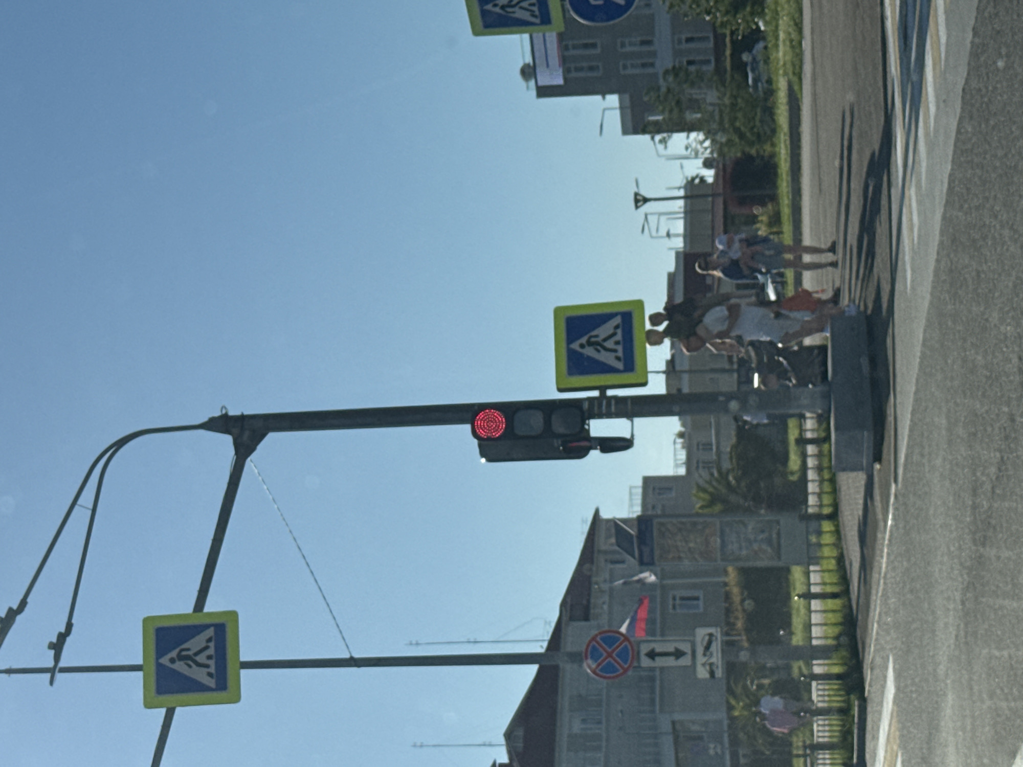 This image, captured in Sirius, Russia, depicts a daytime street scene focused on a pedestrian crossing under a clear blue sky. The original orientation of the image is rotated, with the sky appearing on the left when viewed directly.
In the foreground, a prominent metal pole supports a horizontal traffic light, which is displaying a solid red light, indicating that vehicular traffic has the right-of-way and pedestrians should wait. Attached to this pole, directly beneath the traffic light, is a yellow square pedestrian crossing sign, featuring a white walking figure on a blue background. Another identical pedestrian crossing sign is visible further to the left, mounted higher up on a separate arm.
On the right side of the image, a group of approximately five to seven pedestrians are gathered on the sidewalk, appearing to wait patiently at the edge of the crosswalk. They are dressed in casual, light-colored clothing, suggesting warm and sunny weather. The road in front of them clearly shows the white zebra stripes of the pedestrian crossing.
In the background, several multi-story buildings are visible, interspersed with green trees and hedges. Additional road signage includes a circular sign with a red cross on a blue background (a 'No Stopping' or 'No Waiting' sign), and a white rectangular sign with black arrows pointing left and right, likely indicating traffic directions or a two-way street. The overall scene suggests a typical urban or suburban environment on a bright, sunny day. No discernible text is present on any signs, buildings, or other elements beyond symbols and arrows.