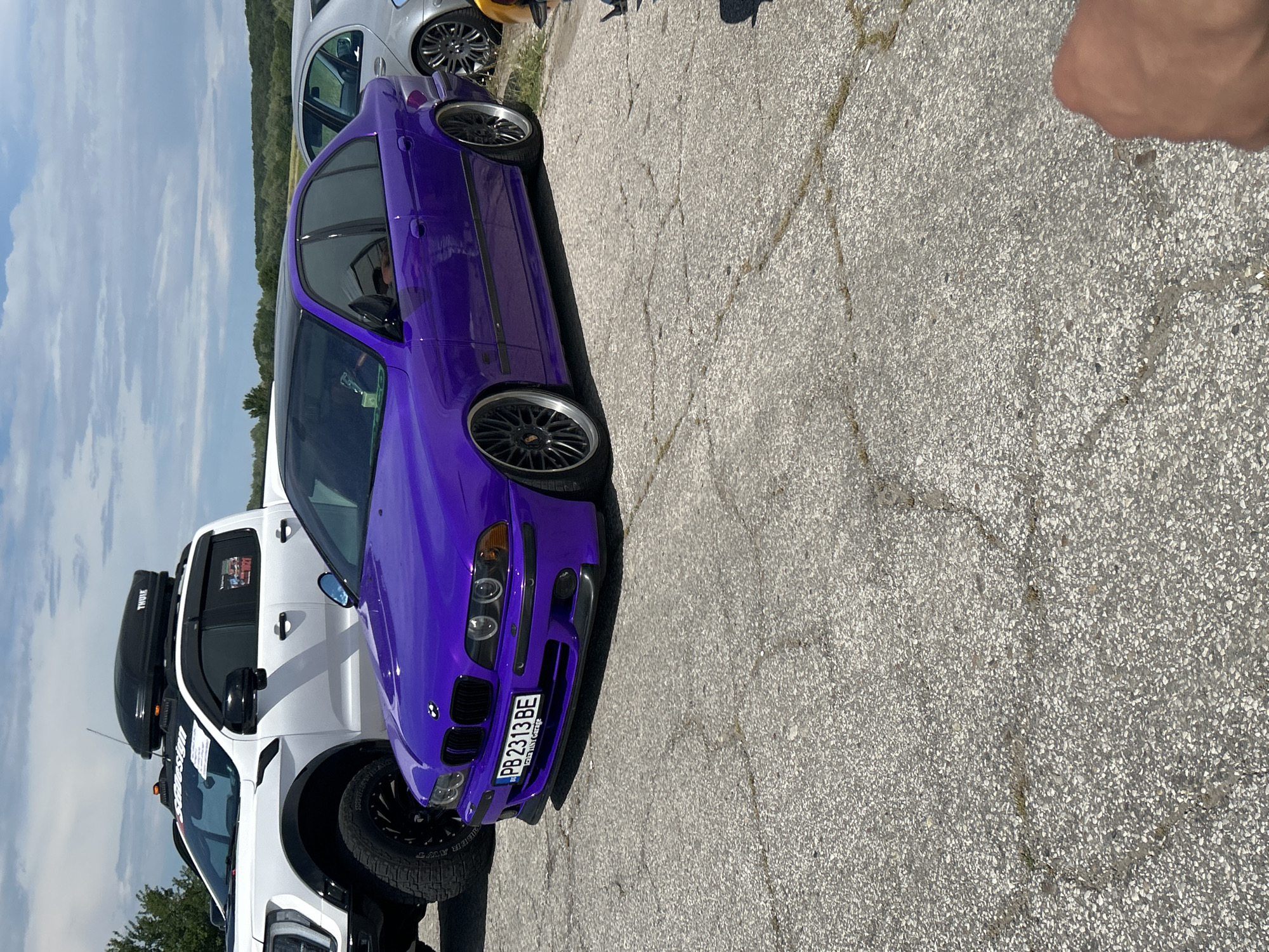 The image shows a purple BMW car parked on an asphalt surface. The car has a Bulgarian license plate "PB 2313 BE". Adjacent to the BMW is a white pickup truck with a black Thule roof box. In the background, another silver car can be partially seen. The location is likely Dalbok Izvor, Bulgaria, based on the license plate information. A human hand is visible on the upper right. The sky is visible in the background.