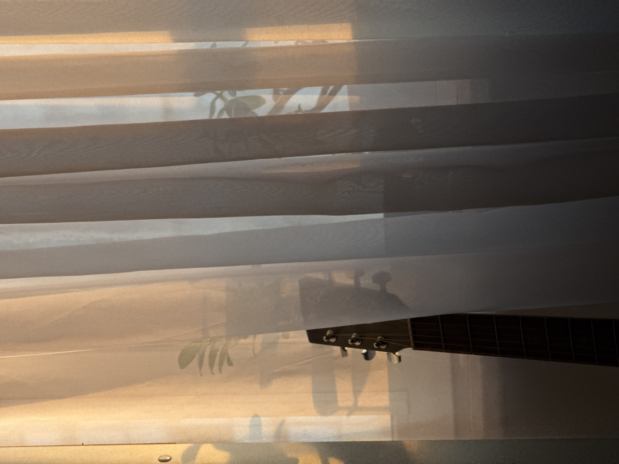 The image is a close-up shot of horizontal blinds that are partially covering a window. Through the translucent fabric of the blinds, a guitar is visible, specifically its headstock and fretboard. The guitar appears to be an acoustic guitar, with tuning pegs and strings clearly discernible. There are also faint reflections of plant leaves, suggesting that there might be plants near the window. The lighting in the image suggests it is either early morning or late afternoon, with a warm, golden hue filtering through the blinds, casting soft shadows and highlights. This warm light indicates a calm and perhaps serene atmosphere. The scene is set indoors, looking out through a window. There is no visible text in the image. The location context provided is Krasnodar, Russia, though there are no specific visual cues in the image to confirm this location.