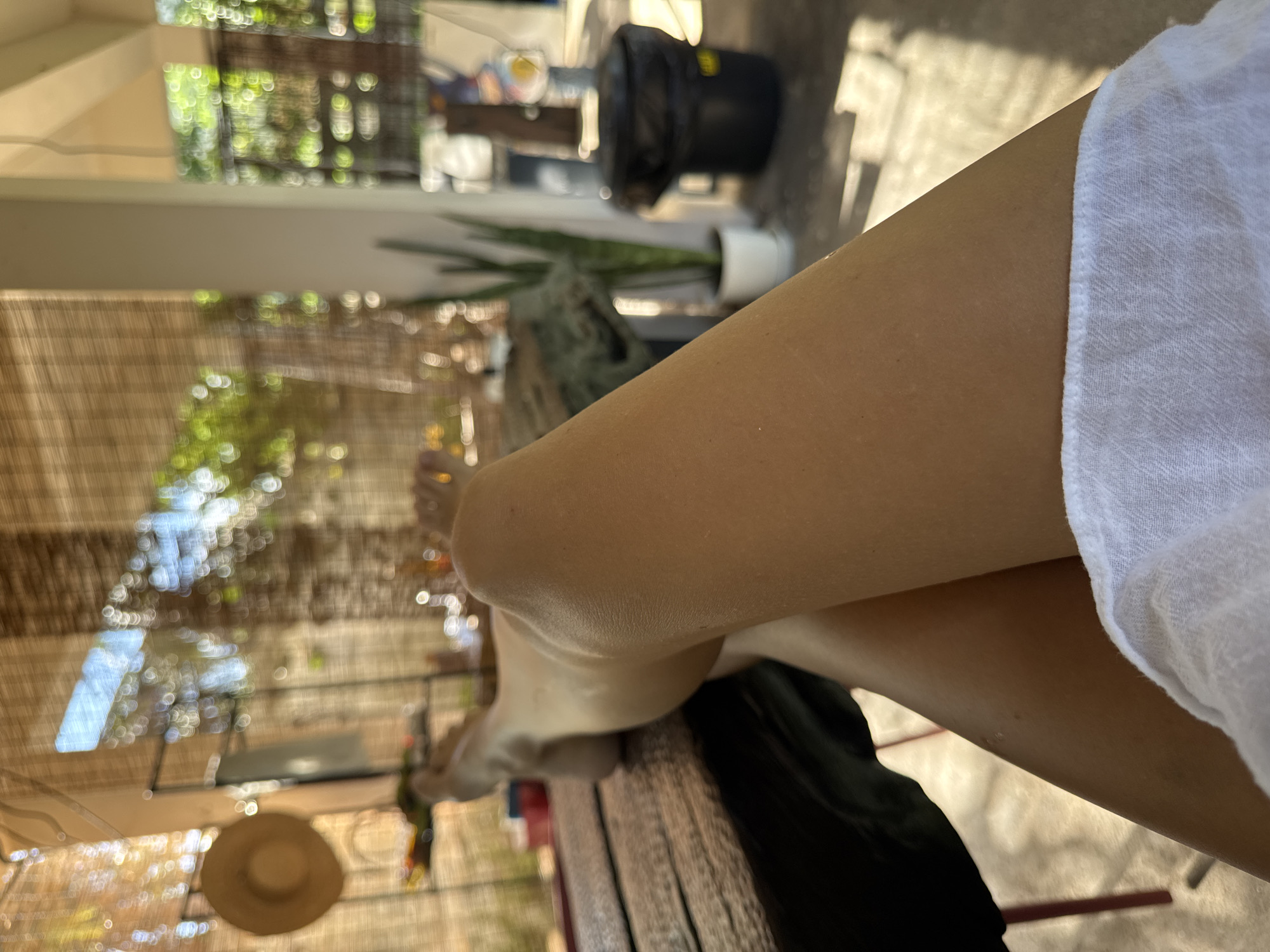 A first-person perspective shot shows a person relaxing outdoors, with their legs crossed and propped up on a wooden surface. The background is a blurred view of a patio or porch area, featuring a bamboo-style screen, some greenery, a black trash can, and a white potted plant. Sunlight filters through the screen, creating dappled light. The person is wearing white fabric covering their lower body. The setting appears to be residential or a relaxed outdoor dining area.