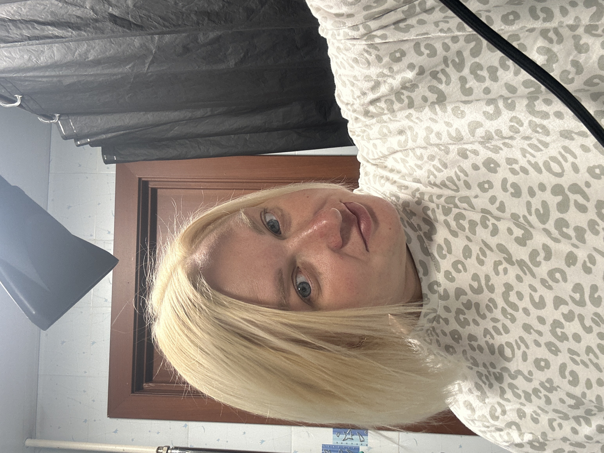 The image is a close-up, vertically oriented selfie of a woman with blonde hair and blue eyes. She is wearing a light-colored sweater with a gray leopard print. The background shows a dark fabric covering on the left, a wooden door frame in the center, and tiled walls with a light blue pattern on the right. A black light fixture is visible on the far left. The lighting suggests it might be indoors, possibly in a bathroom or a room set up for photography. The woman's expression is neutral, and the image appears to be taken in the daytime. There is no visible text in the image.