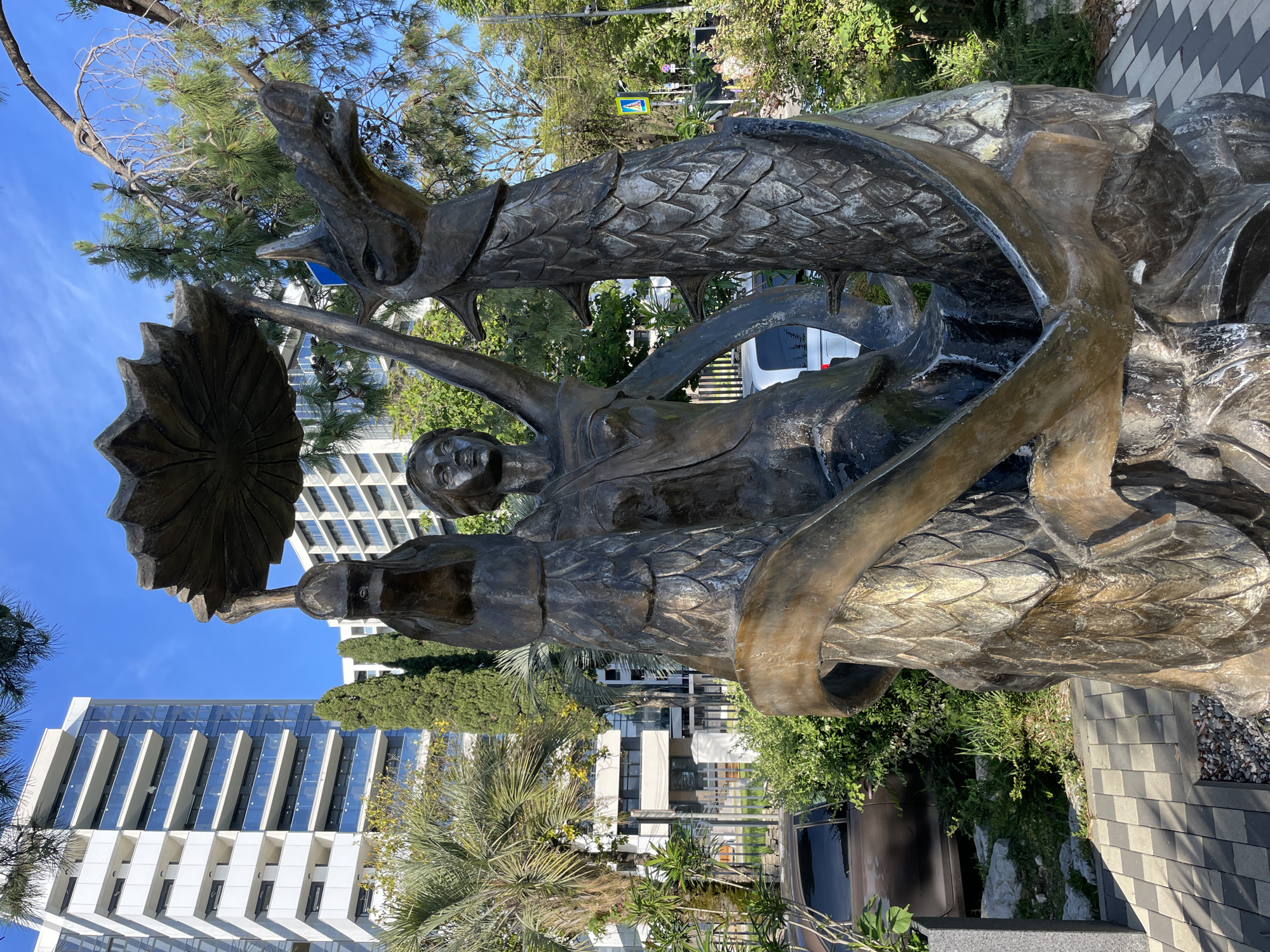 The image depicts a bronze sculpture in Sochi, Russia, featuring a female figure holding a large, lotus-like object above her head, flanked by two dragon-like creatures. The sculpture is situated outdoors, with modern buildings and lush greenery in the background. The buildings are multi-story, white and glass structures, characteristic of contemporary architecture in resort cities. Palm trees and tall, slender cypress trees are visible, suggesting a warm, possibly subtropical climate. The ground around the sculpture is paved with interlocking stones and gravel. Sunlight casts distinct shadows, indicating a clear, sunny day. A brown car is parked to the left of the sculpture, and in the distance, a pedestrian crossing sign and some people can be seen, suggesting a public area. The sculpture itself is detailed, with the dragons exhibiting scaly textures and sharp features, while the female figure has a serene expression. The overall scene combines art and urban landscape in a vibrant setting.