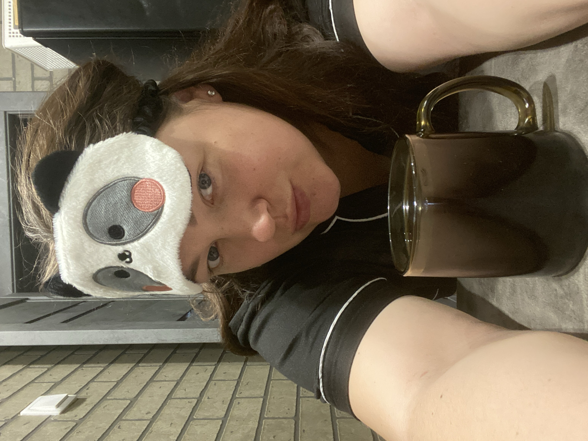A young woman lies on what appears to be a tile floor. She wears a panda-themed eye mask and dark pajamas with white piping. A dark, reflective mug with a light-colored handle sits beside her. The background includes a radiator and what could be the edge of a counter or shelf. The location of Sochi, Russia, cannot be determined from the image.