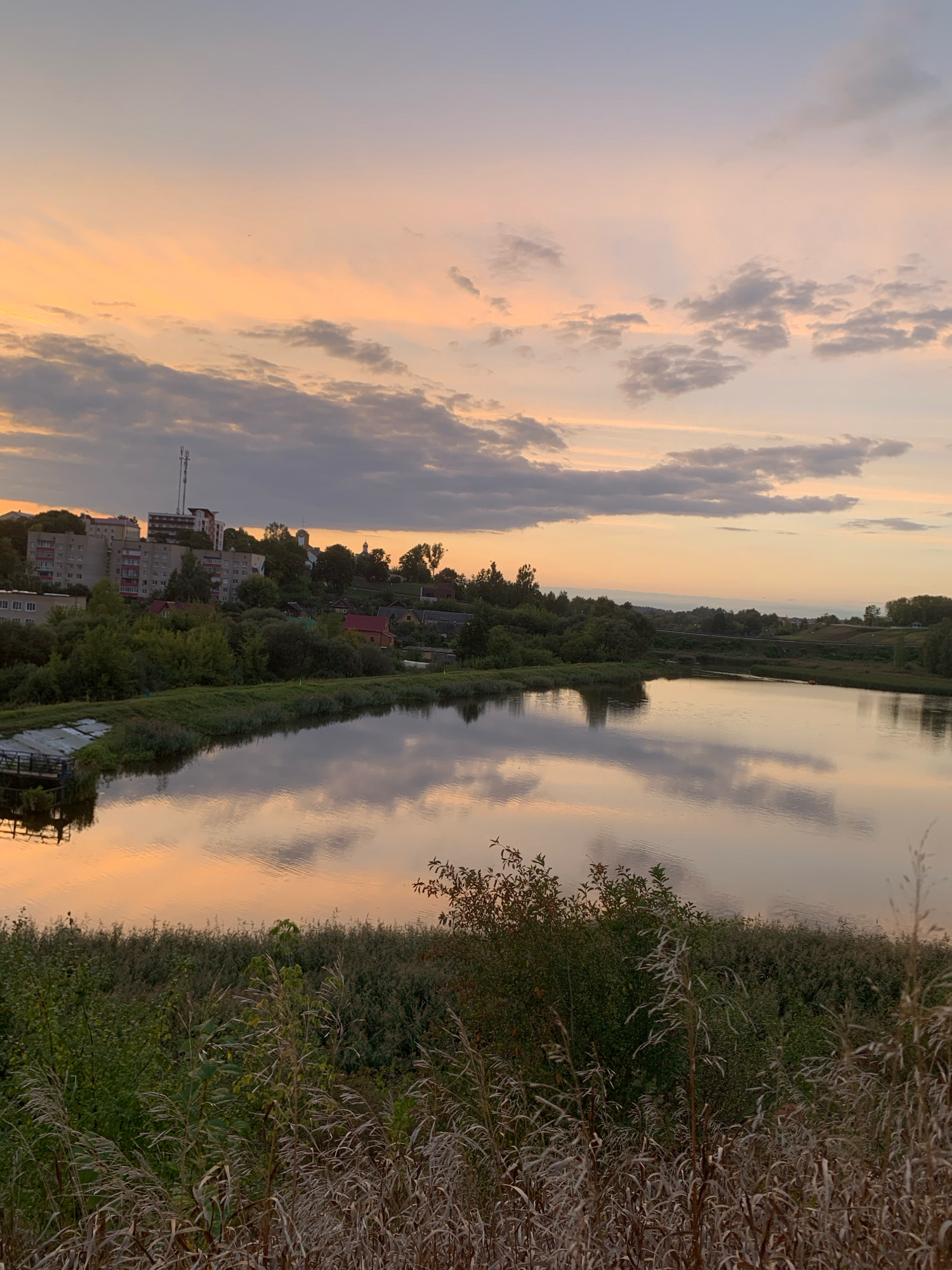 The image depicts a tranquil lakeside scene in the city of Kapyĺ, Belarus, during sunset. The sky is painted with hues of orange and pink, with scattered clouds reflecting on the calm water. The foreground is dominated by dry grass and reeds, creating a textured contrast with the smooth water surface. On the far bank, a small cluster of buildings, including apartment blocks and houses, is visible, nestled amongst lush green trees. A small structure, possibly a pier or dock, extends into the water on the left side of the frame. The overall atmosphere is serene and peaceful, capturing the essence of a quiet evening in the Belarusian countryside.