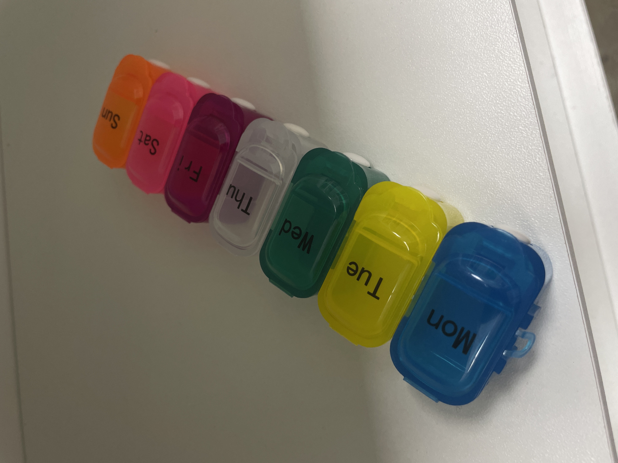 A stack of seven pill organizers, each a different color, are lined up diagonally on a white shelf. The pill organizers are labeled with the days of the week in black text. From bottom to top, the organizers are blue labeled "Mon", yellow labeled "Tue", green labeled "Wed", clear labeled "Thu", purple labeled "Fri", pink labeled "Sat", and orange labeled "Sun". The organizers are all closed. The background is a plain white wall. The image is taken from a slightly elevated angle, looking down at the pill organizers.