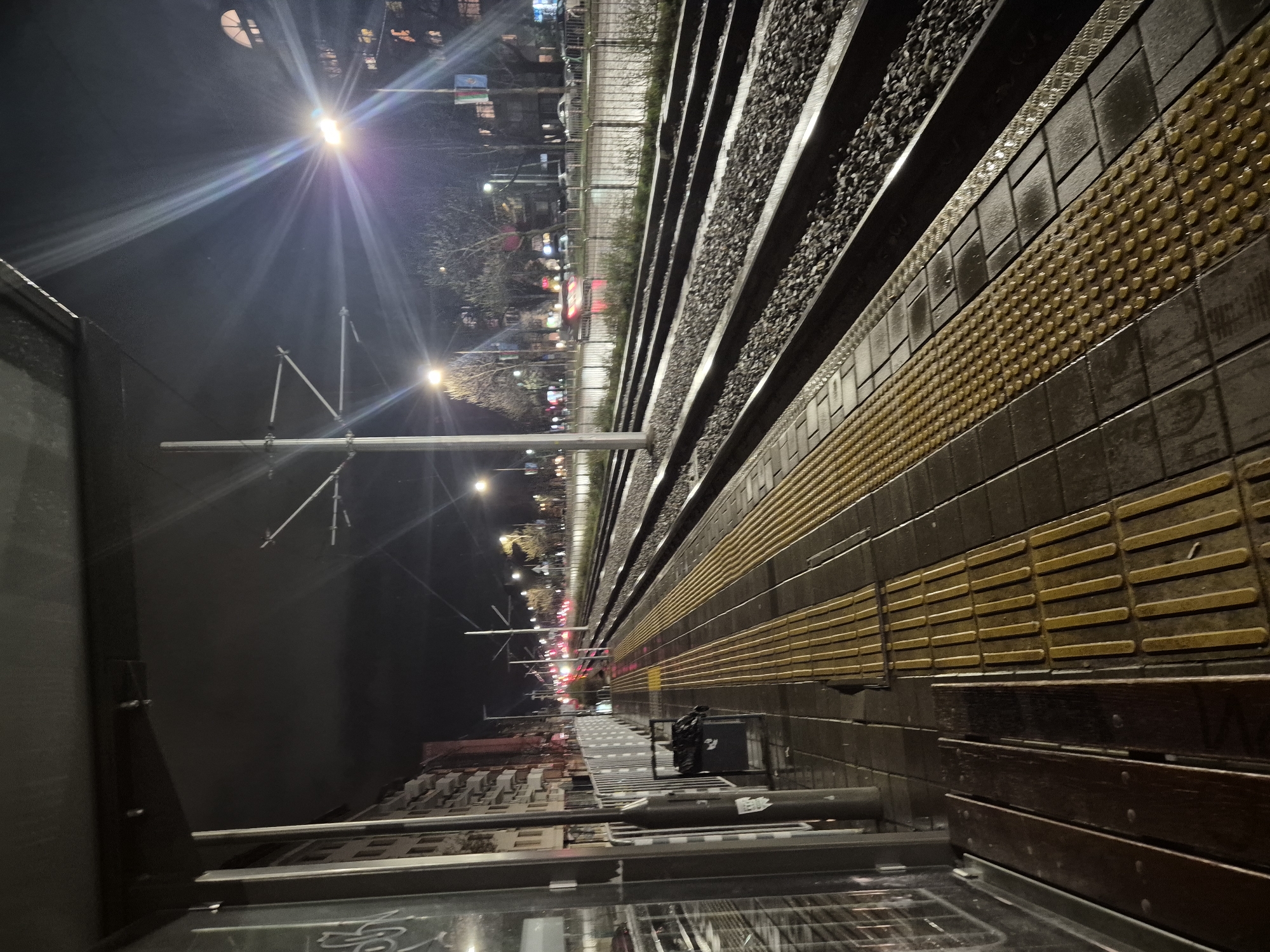 The image depicts a nighttime view of what appears to be a train platform in Sofia, Bulgaria. The angle is low and slightly tilted. The platform is lined with tactile paving, and a row of dark wooden benches. Parallel to the platform are train tracks filled with ballast. Beyond the tracks, a city street can be seen with street lights and blurred vehicle lights. Overhead electrical wires and support structures are visible extending over the tracks and platform. The foreground suggests the view is from a glassed-in waiting area.