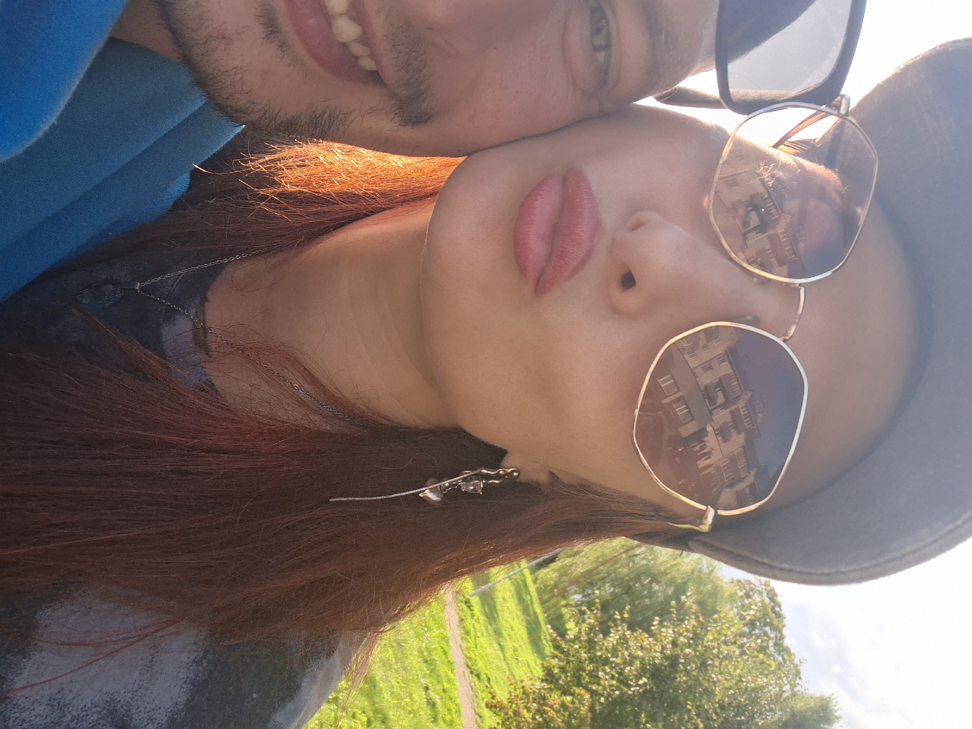 This is a close-up, outdoor, eye-level, selfie-style photograph of two individuals. The person in the foreground, on the right, has reddish-brown hair, is wearing large, hexagonal-shaped sunglasses with reflective lenses, a grey cap, and a silver earring with a heart charm. Their lips are parted as if blowing a kiss. The reflection in their sunglasses shows a multi-story building with balconies. To the left of the person on the right is another individual, only partially visible, showing the left side of their face and part of their blue jacket. This individual has light-colored eyes and is smiling, revealing teeth. The background suggests an outdoor setting with green grass and trees, illuminated by sunlight. The scene appears to be in a park or grassy area, possibly in the city of Myadzyel, Belarus, given the context provided.