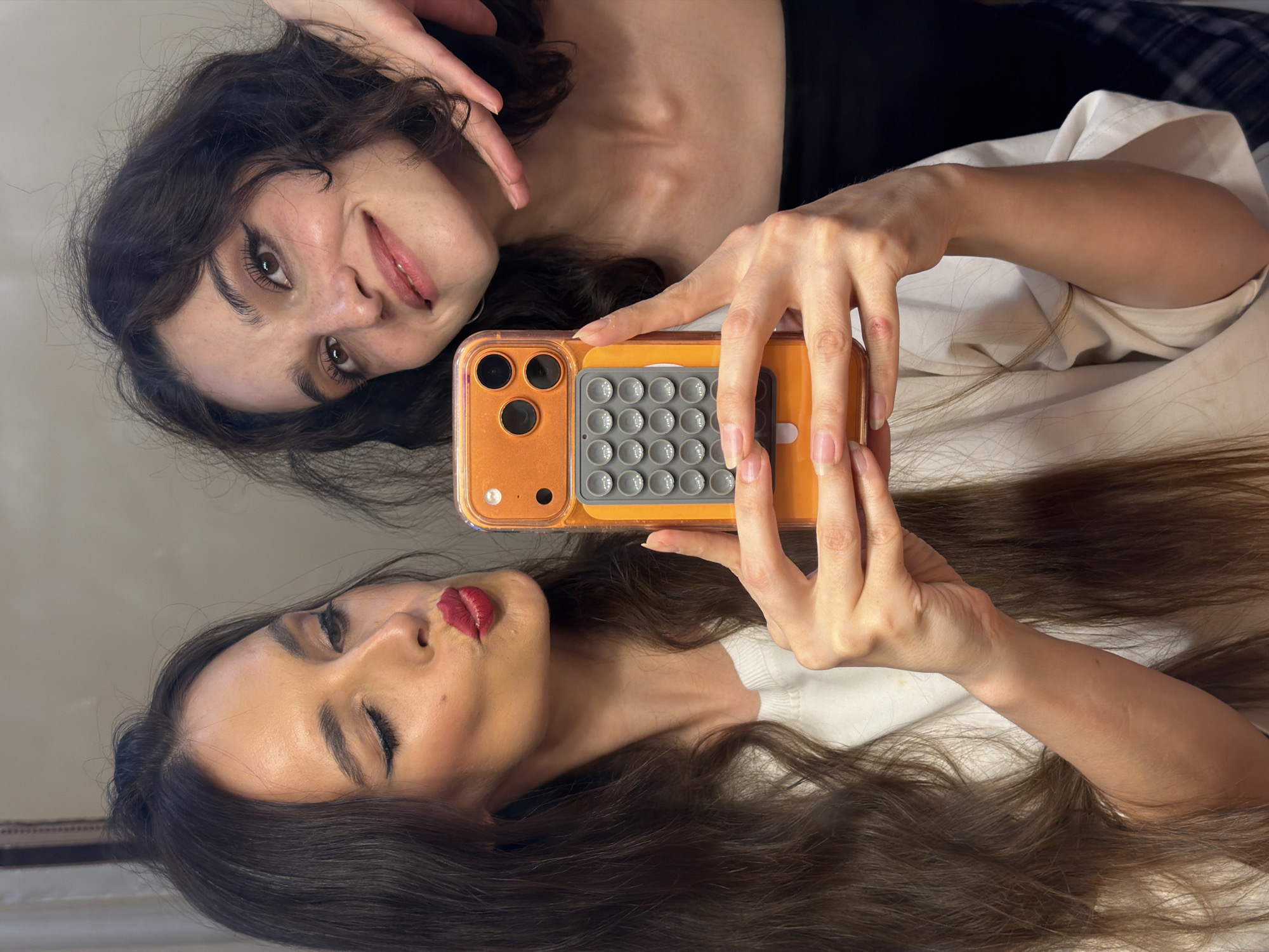 This is a waist-up, medium shot selfie taken by two young women indoors. The woman on the left is leaning forward, puckering her lips and holding a bright orange phone case in her left hand. She has long, dark brown hair, winged eyeliner, and red lipstick. She is wearing a white long-sleeved shirt. The woman on the right is smiling and leaning her head and left arm on the shoulder of the woman on the left, with her right hand near her chin. She has dark, wavy hair and is wearing a black top. The background appears to be a mirror reflecting the scene. There are no identifiable landmarks suggesting the location is in Ufa, Russia.