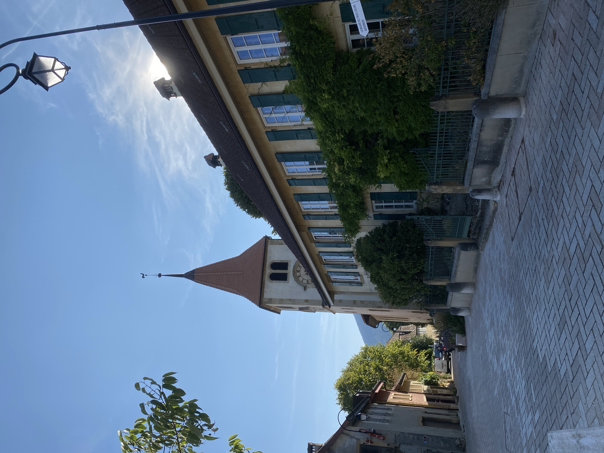 This is a medium shot, eye-level, outdoor, daytime photograph taken in the town of Milvignes, Switzerland. The image depicts a cobblestone street leading uphill towards a building with a distinctive bell tower. Sunlight streams through the sky, creating lens flare and illuminating the scene. The street is lined with buildings, some of which are adorned with green shutters and climbing ivy. A lamppost with a decorative lantern stands on the right side of the street. A signpost is visible on the right side of the building, with text that is too small to read. A tree with green leaves is on the left side of the frame, partially obscuring the view. The sky is blue with some wispy clouds. In the distance, mountains are visible. A few vehicles are parked along the street. The overall impression is one of a quaint, historic European village.