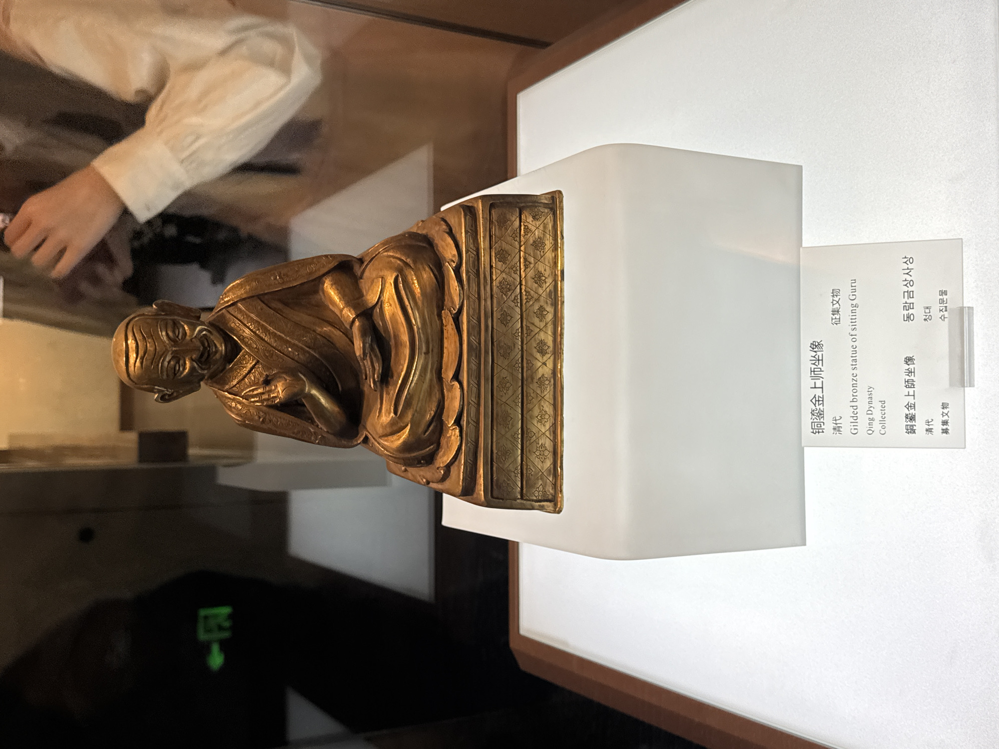 A photograph showcases a gilded bronze statue of a seated guru, likely from the Qing Dynasty, situated in Dunhuang City, China. The statue rests on a white pedestal with accompanying text in Chinese and Korean, identifying it as a "Gilded bronze statue of sitting Guru, Qing Dynasty, Collected." A partially visible person in a white shirt is reflected in the glass case surrounding the display. The environment is dimly lit, further accentuating the gold tone of the statue.