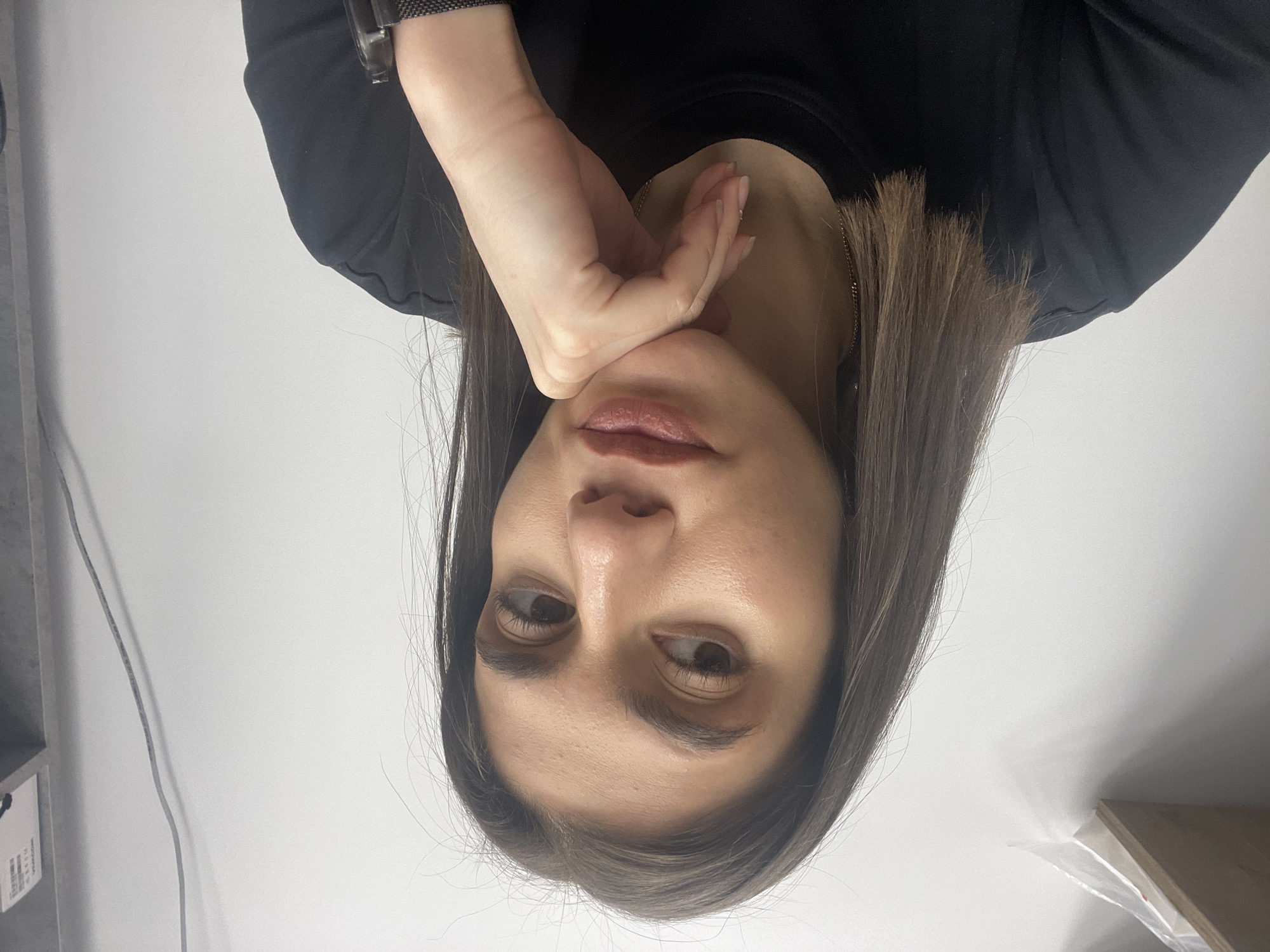 A woman with dark hair and eyes poses with her hand near her face. She wears a black shirt and a watch. The background is a plain white wall. The location is unspecified and there is no indication that it is Sofia, Bulgaria.