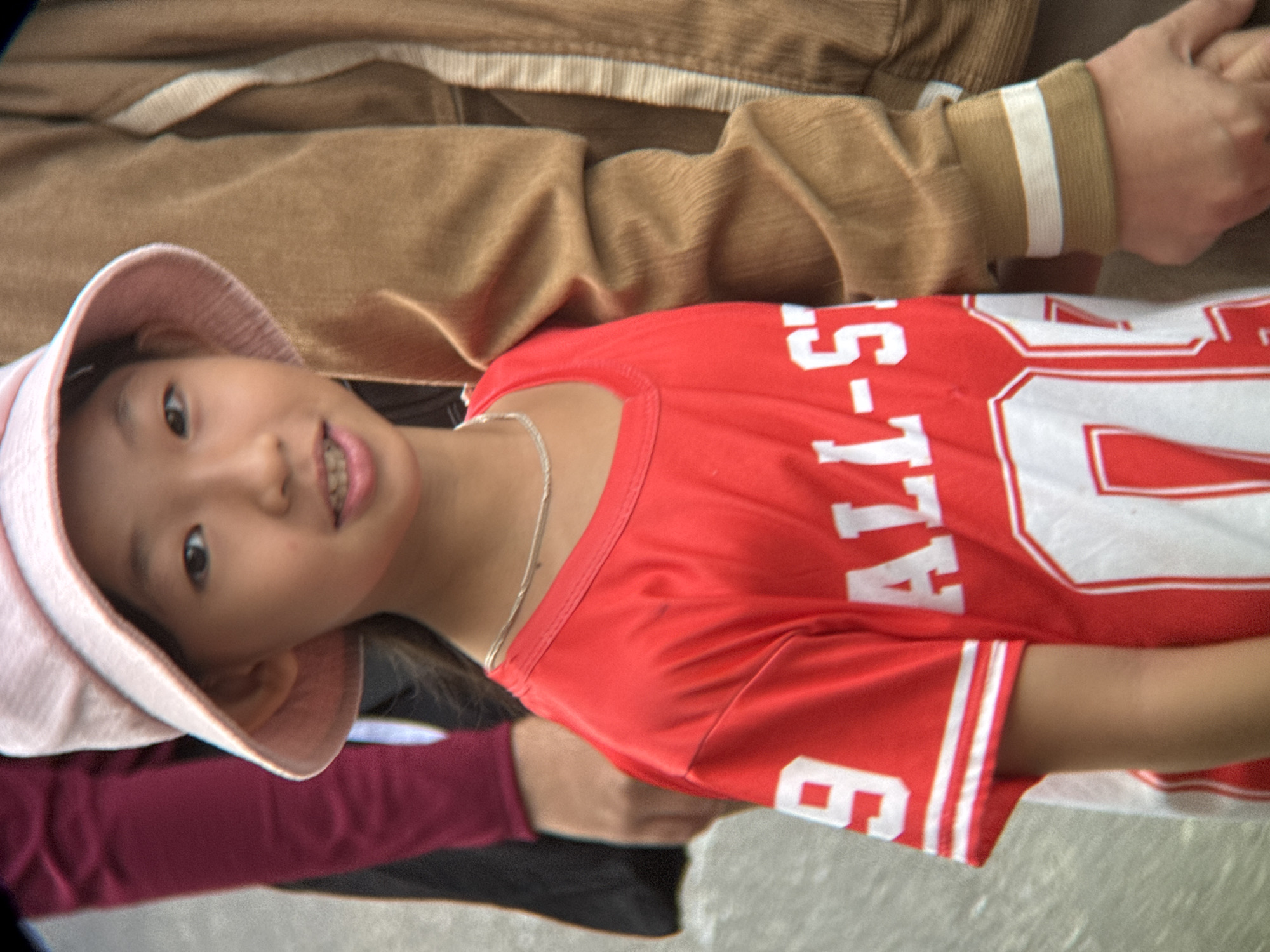 A young Asian girl wearing a pink bucket hat and a red t-shirt with white lettering stands in front of an adult wearing a tan jacket. The girl has a silver necklace around her neck. The t-shirt has the number "9" on the left sleeve and white lettering that reads "ALL-ST" on the front. The adult's jacket has white stripes on the cuffs. They are outdoors, with a textured, greyish-green surface in the background. The image appears to have been taken on a street in Nha Trang, Vietnam, as indicated by the surrounding environment and the demographics of the people present.