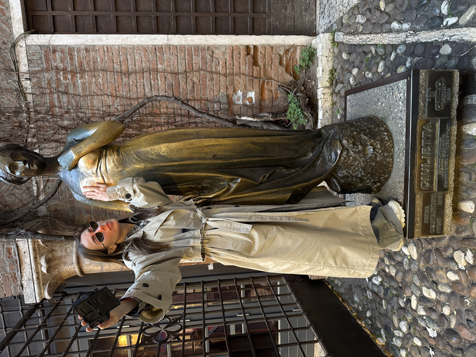 The image depicts a woman interacting with the bronze statue of Giulietta (Juliet) within the courtyard of the Casa di Giulietta in Verona, Italy.
The subject is a female with long brown hair, wearing a beige trench coat over a plaid shirt, gray pants, and gray sneakers. She wears narrow, black oval sunglasses. Her left arm is extended forward, holding a black compact camera or smartphone, pointed towards herself, indicating a selfie action. Her right hand is placed flat against the chest area of the bronze statue to her right. She gazes towards the camera/device.
The bronze statue, approximately life-sized, portrays a female figure in a long, flowing dress, with her left hand resting on her chest. The right breast of the statue exhibits significant wear and a polished, lighter appearance, distinct from the surrounding patina. The statue stands on a dark stone plinth, which in turn rests on a larger rectangular base. A bronze plaque affixed to the front of this base clearly displays "GIULIETTA" in prominent lettering, along with smaller inscriptions "NERO COSTANTINI REGIT 1969" on the left and "DONO DI CATTOLICA 2014" on the right.
The environment features a ground covered in rounded cobblestones. Behind the statue and the woman is a reddish-brown brick wall, showing signs of age and mortar degradation in places. Sparse green vegetation and bare climbing vines are visible against the wall. To the far left, a dark metal gate or railing with decorative elements is partially visible. The lighting is diffused, suggesting an overcast day or shaded area, with no harsh shadows present.